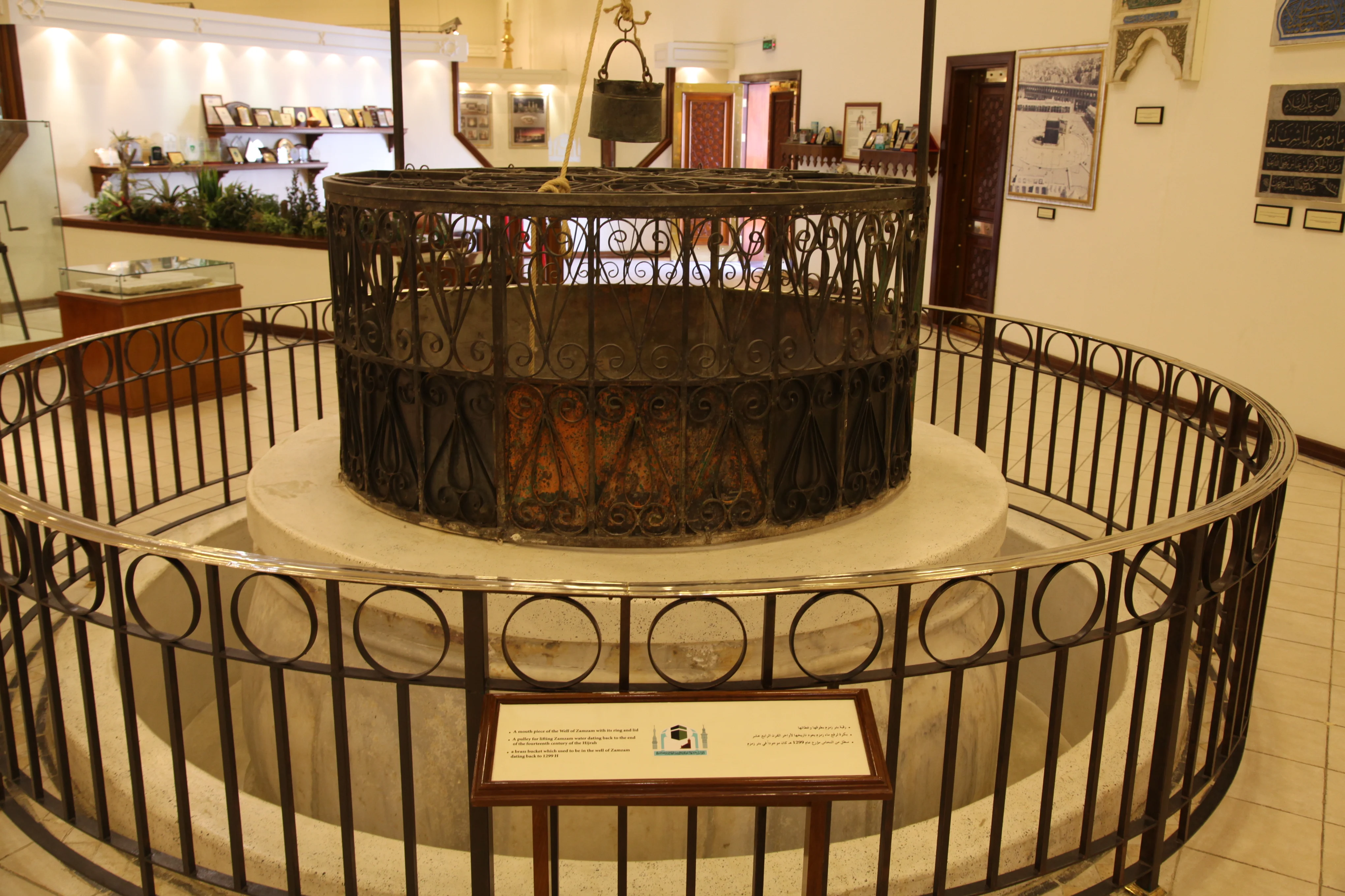 A circular openwork iron well-head with a lid, standing on a low stone plinth inside a museum gallery and enclosed by railings, with a brass bucket suspended above it on a rope from a pulley, and display cases, framed photographs and inscribed panels along the walls behind