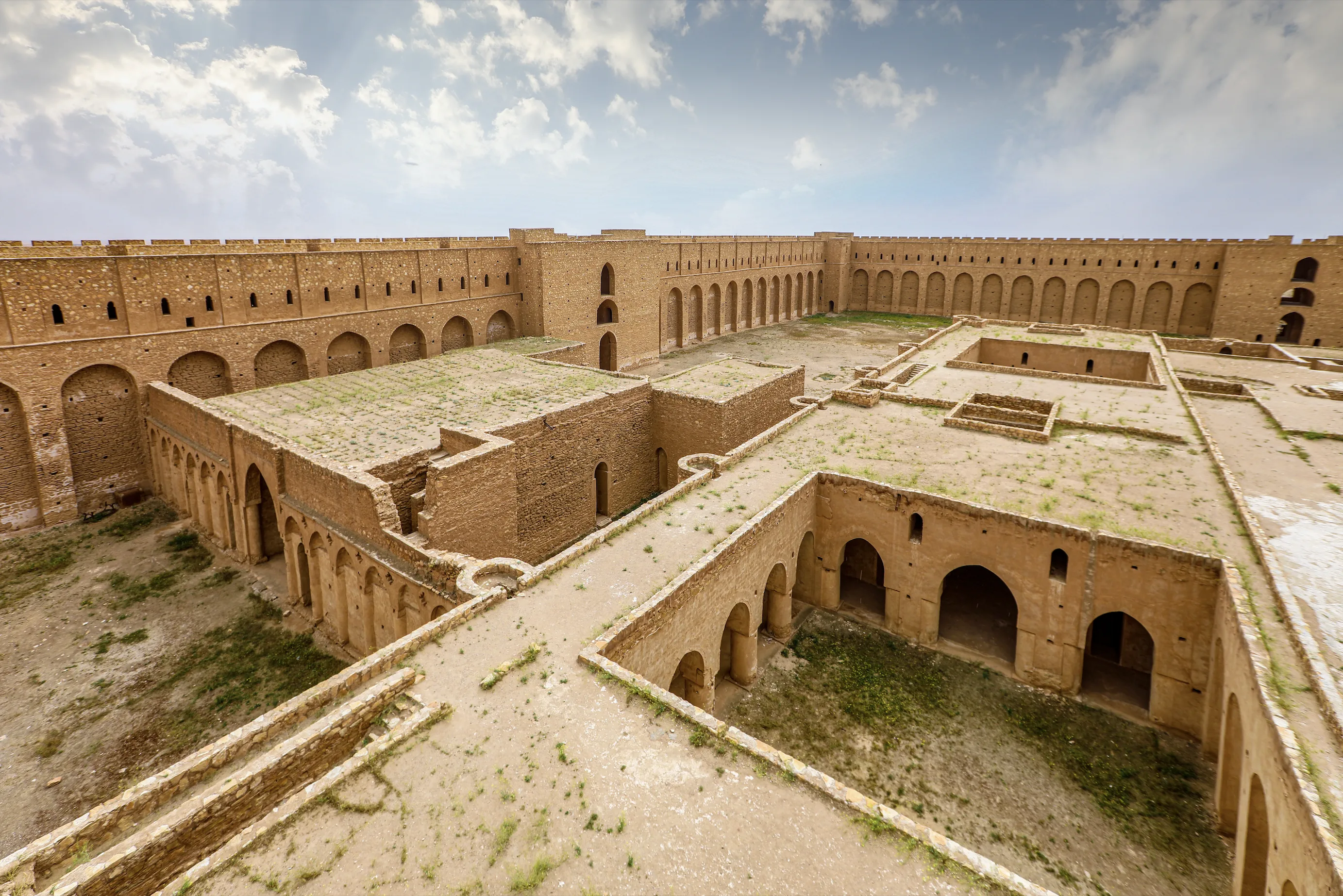 The mud-brick ruins of a large rectangular fortress, its high outer walls lined with arcaded galleries enclosing a courtyard of roofless vaulted chambers, under a cloudy sky
