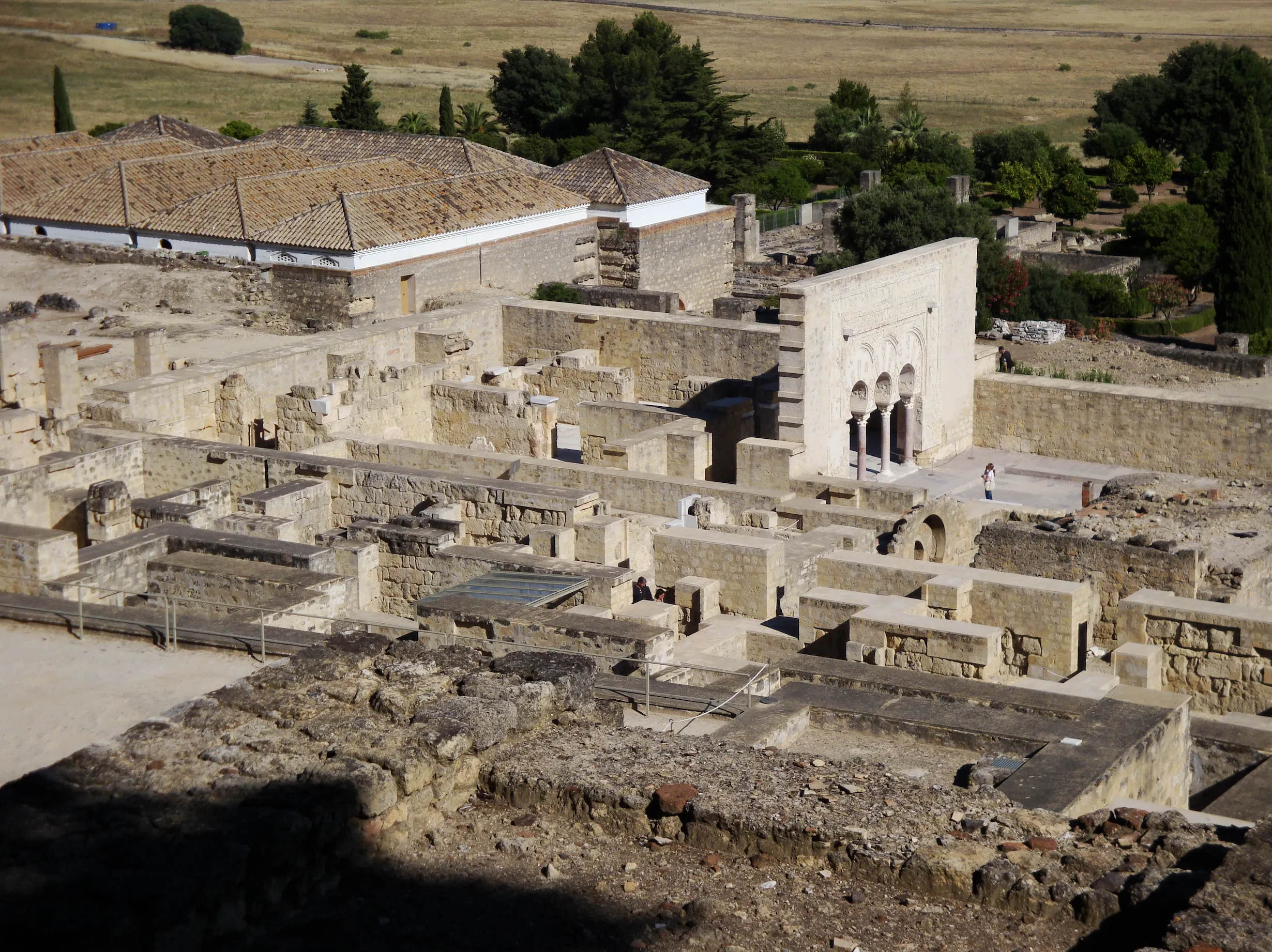 An aerial view of an excavated palace-city, with stone foundation walls, a reconstructed arcaded portico of horseshoe arches on marble columns, and fields and trees beyond