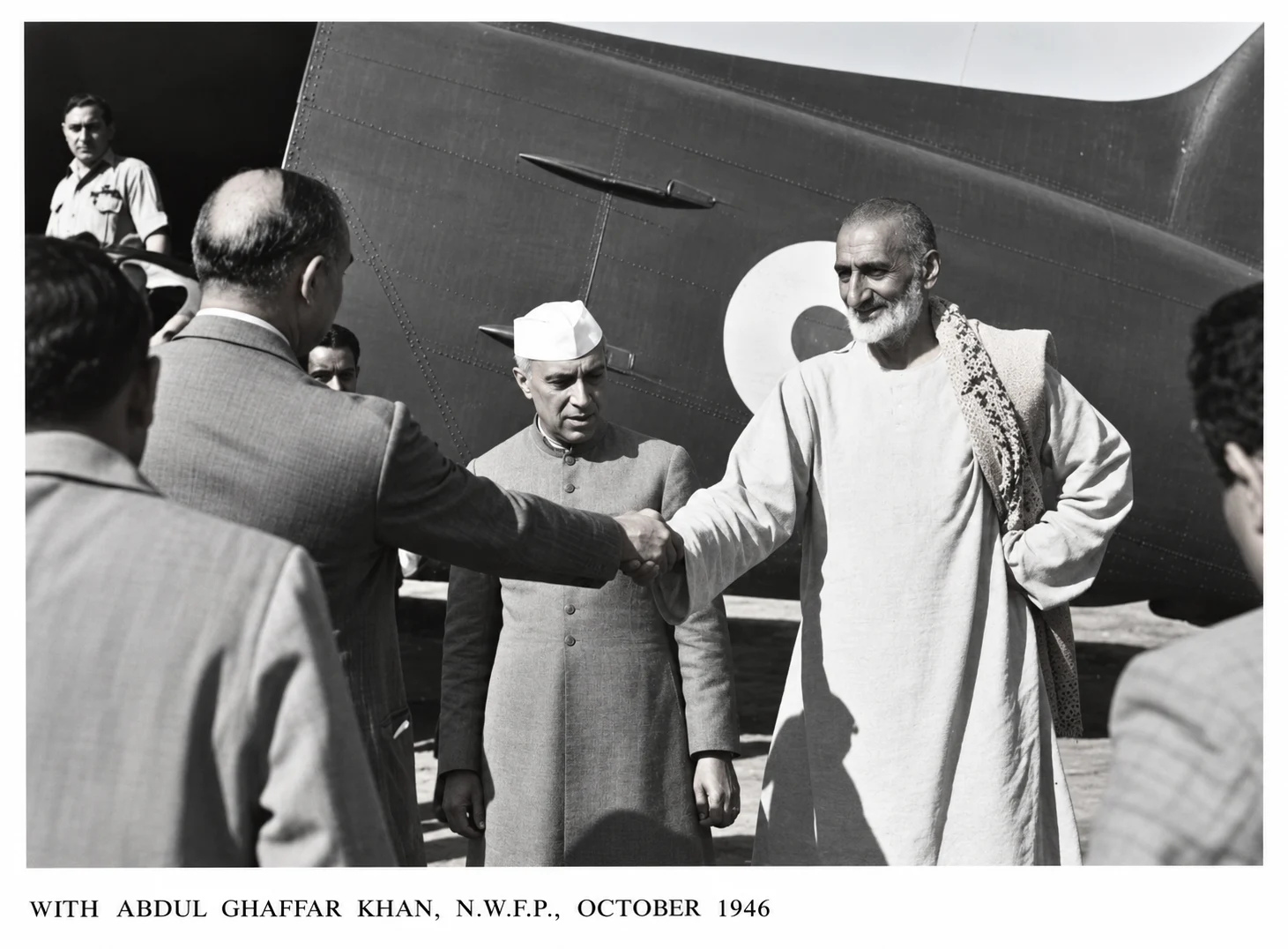 Black-and-white photograph of a tall bearded man in a light ankle-length kurta with a patterned shawl over one shoulder, shaking hands with a man in a suit seen from behind, while a second man in a white cap and buttoned achkan stands between them; the fuselage and wing of an aircraft fill the background