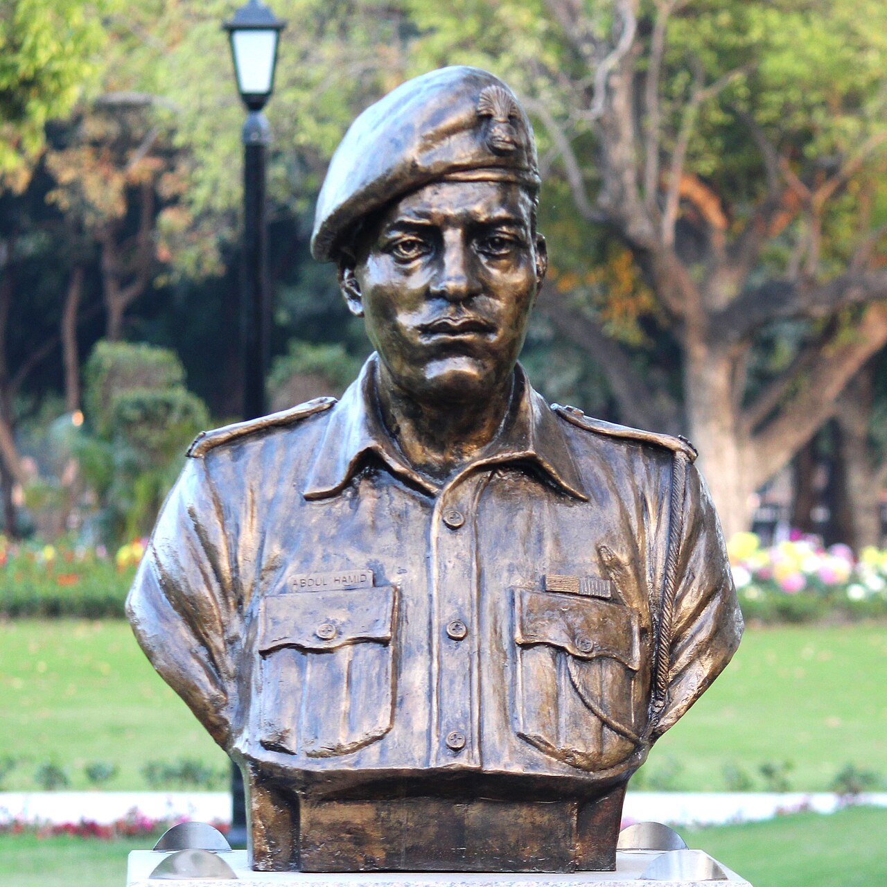 Colour photograph of a bronze bust of Abdul Hamid in army uniform, wearing a beret with a regimental cap badge and a shirt bearing a nameplate reading ABDUL HAMID, mounted on a stone plinth in a garden