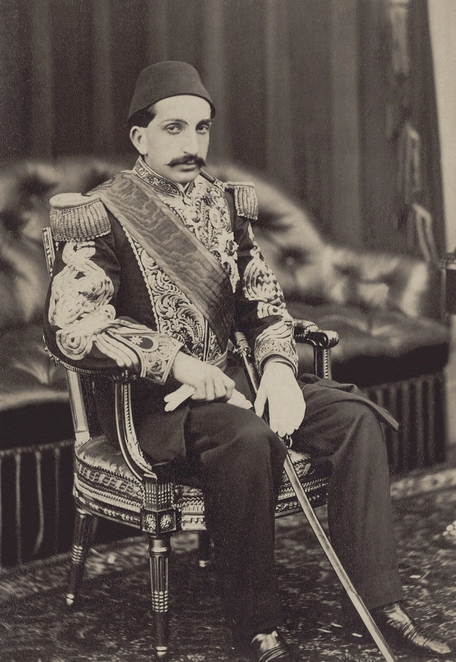 A monochrome studio photograph of a seated man with a moustache wearing a fez and an elaborately embroidered dark court uniform with epaulettes and a sash, one hand resting on a sword