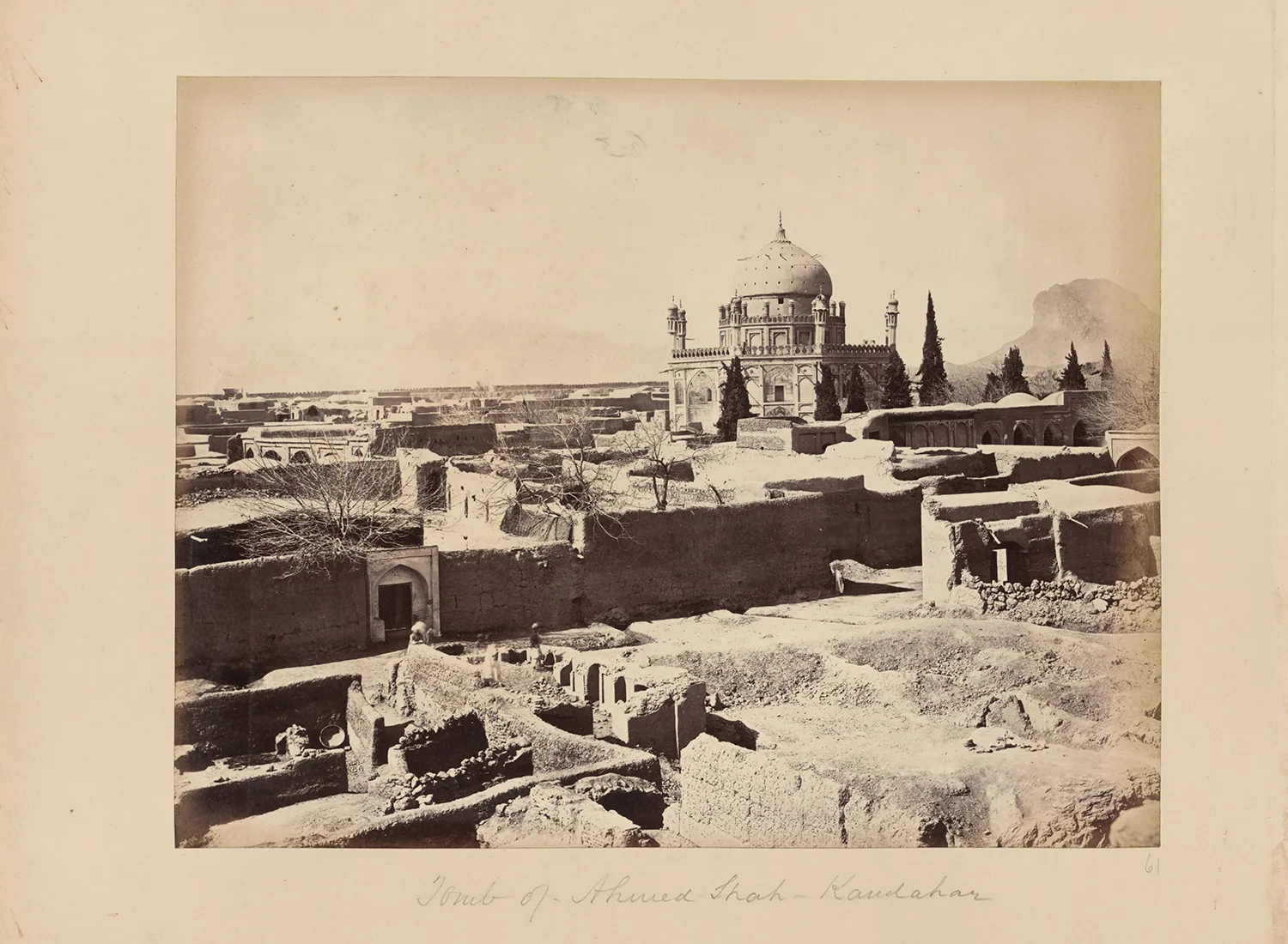 An old sepia-toned photograph of a domed mausoleum with a tiled facade rising above ruined mudbrick walls and cypress trees, with hills in the background