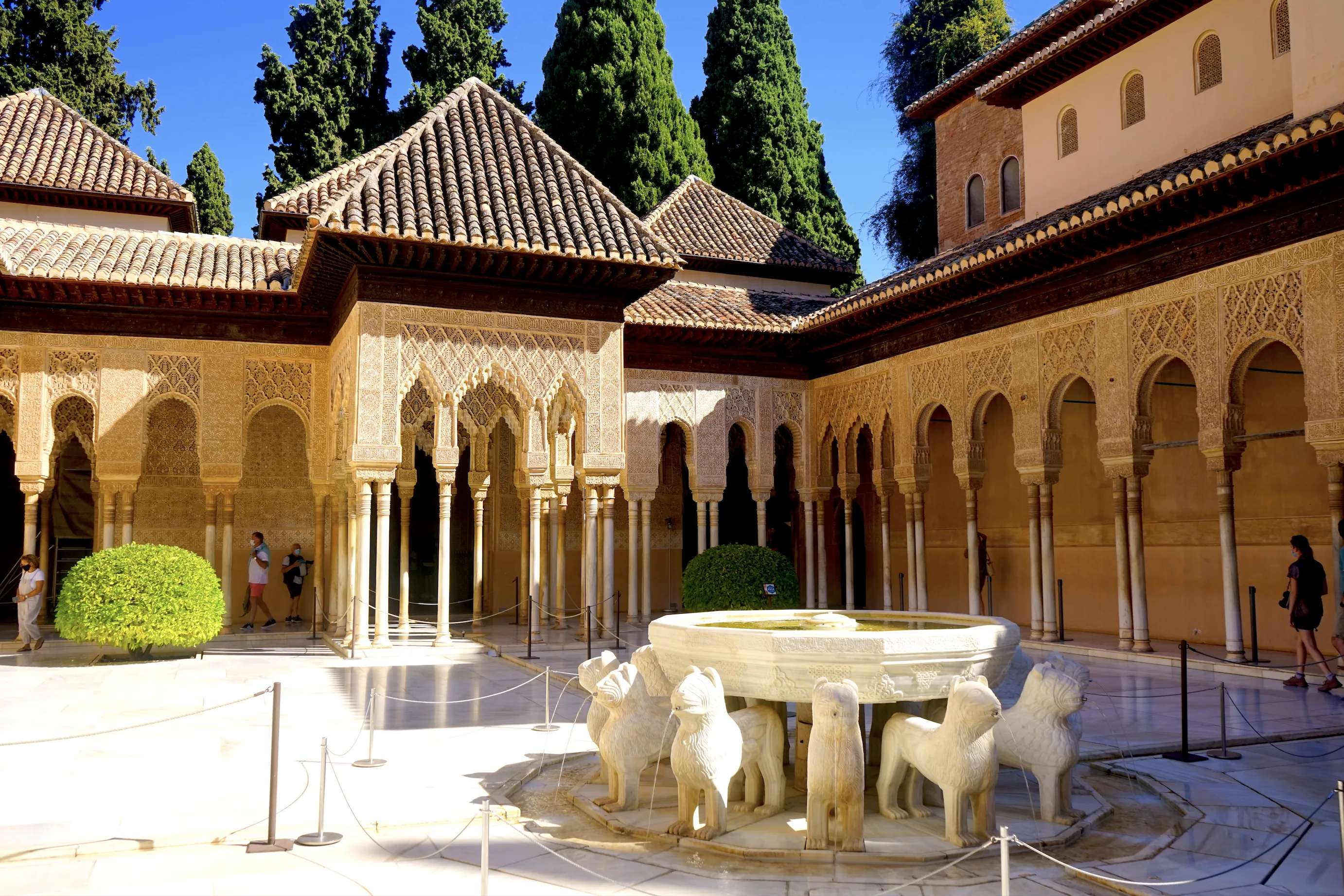 An arcaded courtyard of horseshoe and multifoil arches on slender marble columns with elaborate stucco carving, tiled pyramidal roofs and cypress trees behind, and a marble fountain supported by stone lions in the foreground