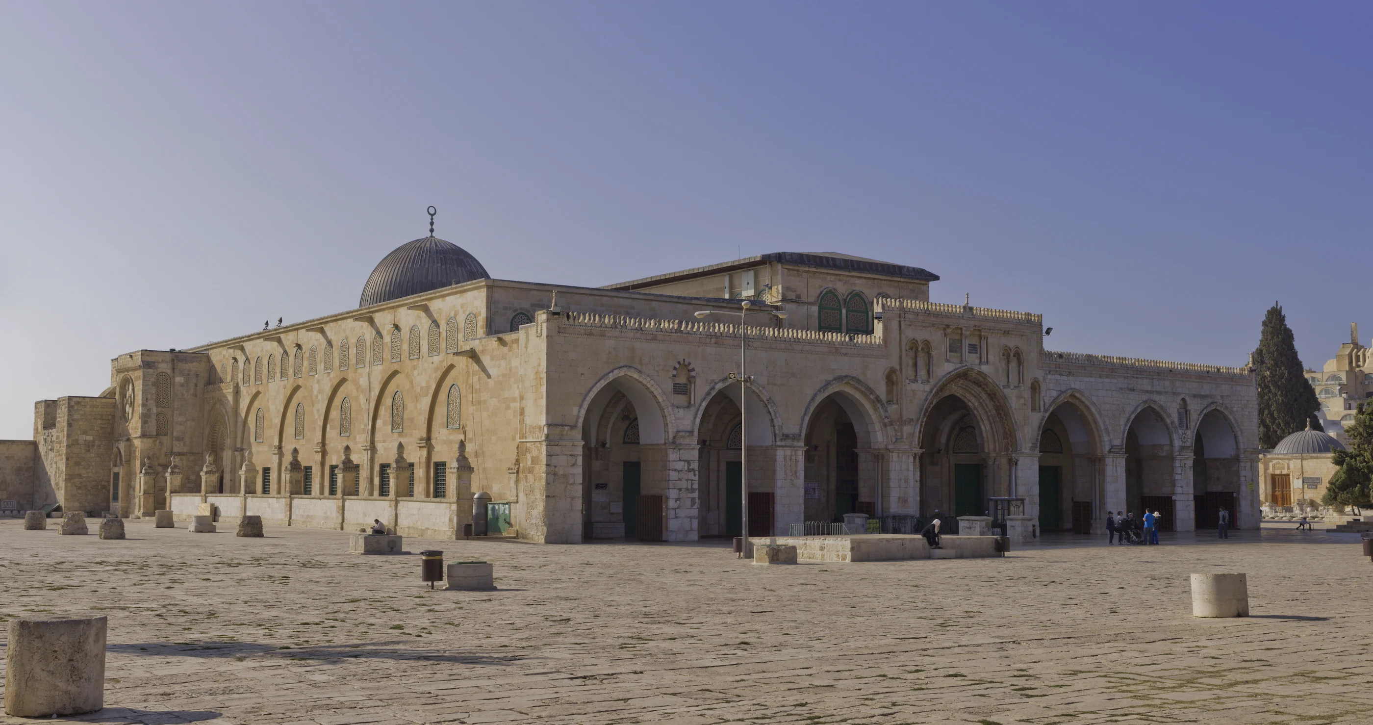 The Al-Aqsa Mosque on the Haram al-Sharif in Jerusalem, a long stone building with a silver-grey lead dome and an arched façade, seen across the paved plaza