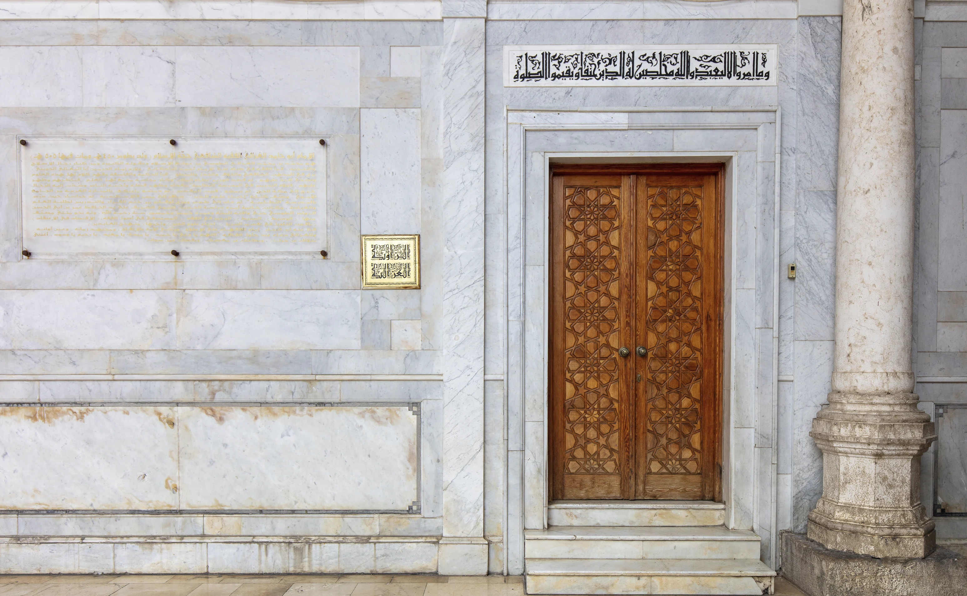 A carved wooden double door with a geometric star-pattern inlay, set in a marble wall beneath a Qur'anic inscription band, with an inscribed marble plaque to its left