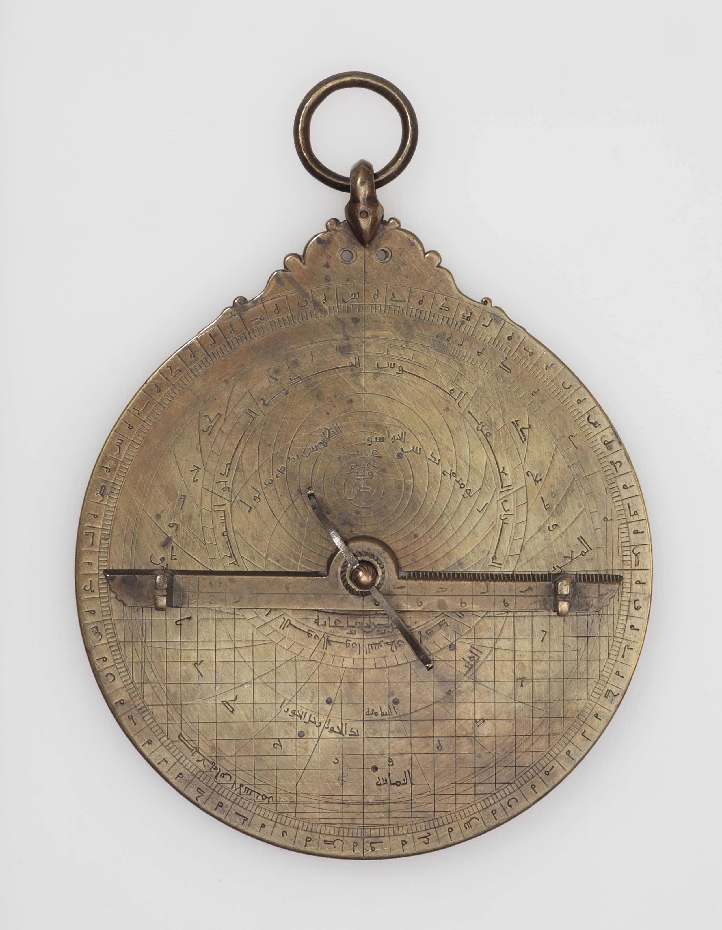 A circular brass planispheric astrolabe with an engraved suspension ring, a pierced rete of pointers over an engraved plate, and a sighting rule across the front, covered in Arabic inscriptions and scales