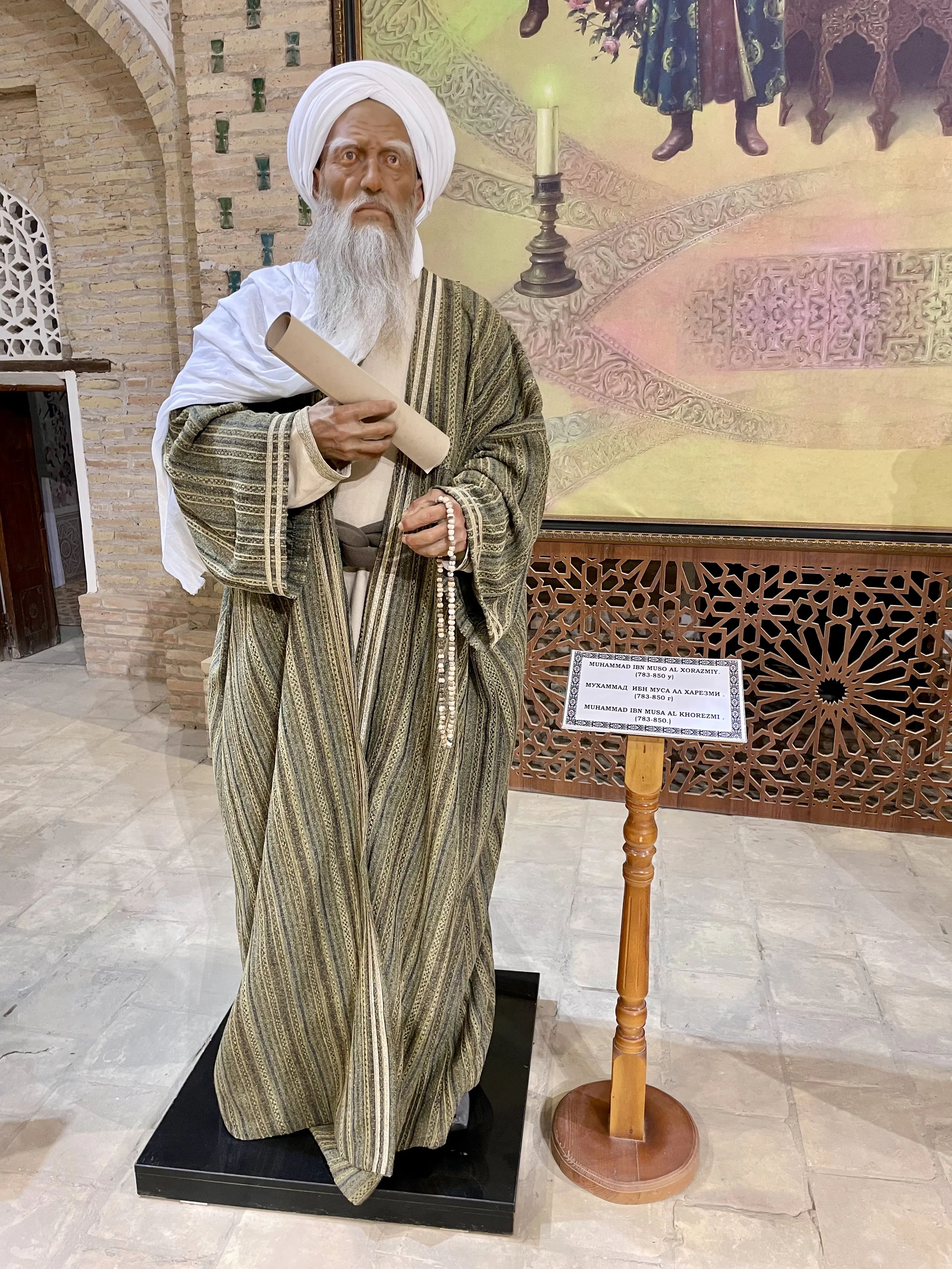 A life-size costumed figure of an elderly bearded scholar in a white turban and shawl over a striped green and gold robe, a rolled scroll tucked under one arm and a string of prayer beads in the other hand, standing on a low plinth in a brick-walled hall beside a wooden lectern carrying a name label in three scripts
