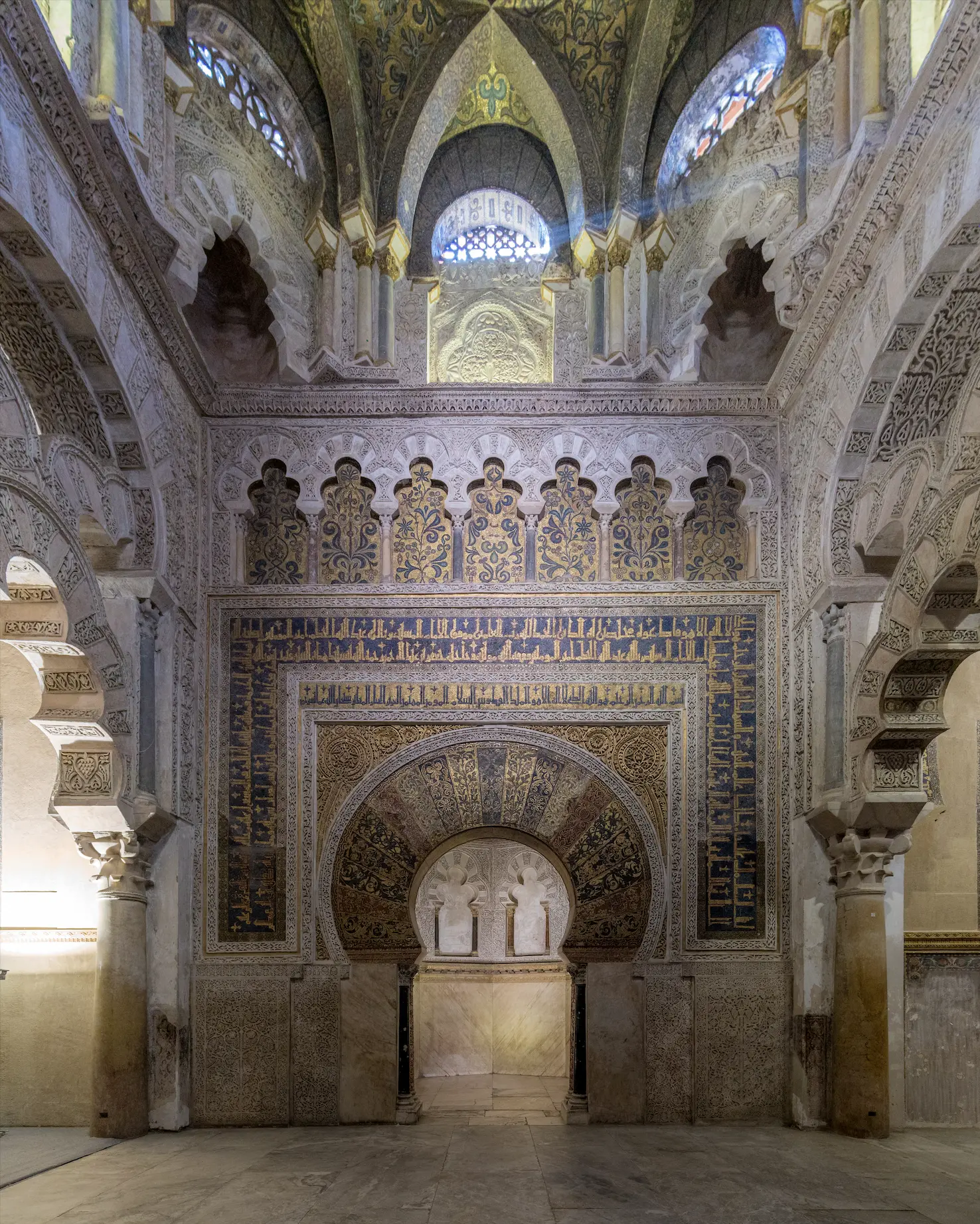 The mihrab of the Great Mosque of Cordoba: a horseshoe-arched niche on marble columns, framed by a band of gold-ground Kufic inscription, with an arcade of seven lobed arches in blue and gold mosaic above it and a ribbed, mosaic-lined vault overhead, flanked by the tall cusped arches of the surrounding chamber