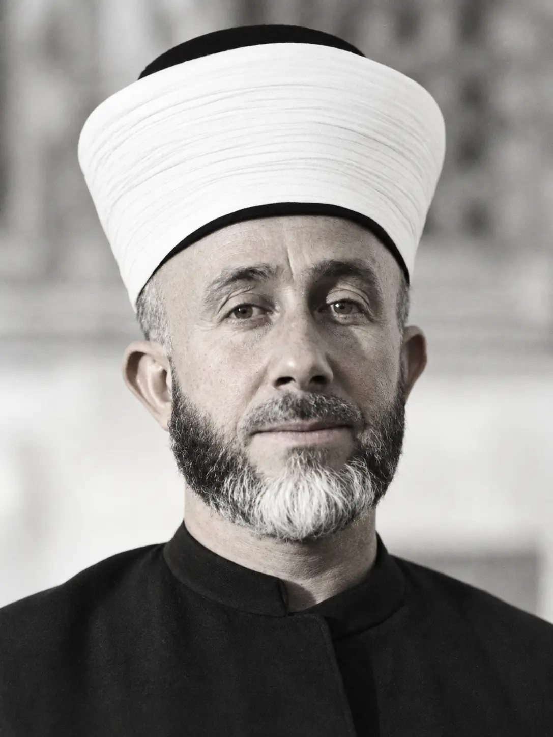 AI-generated impression: A monochrome portrait of a bearded man in a tall white turban wound over a dark cap, wearing a dark high-collared robe, before a blurred background of carved stonework