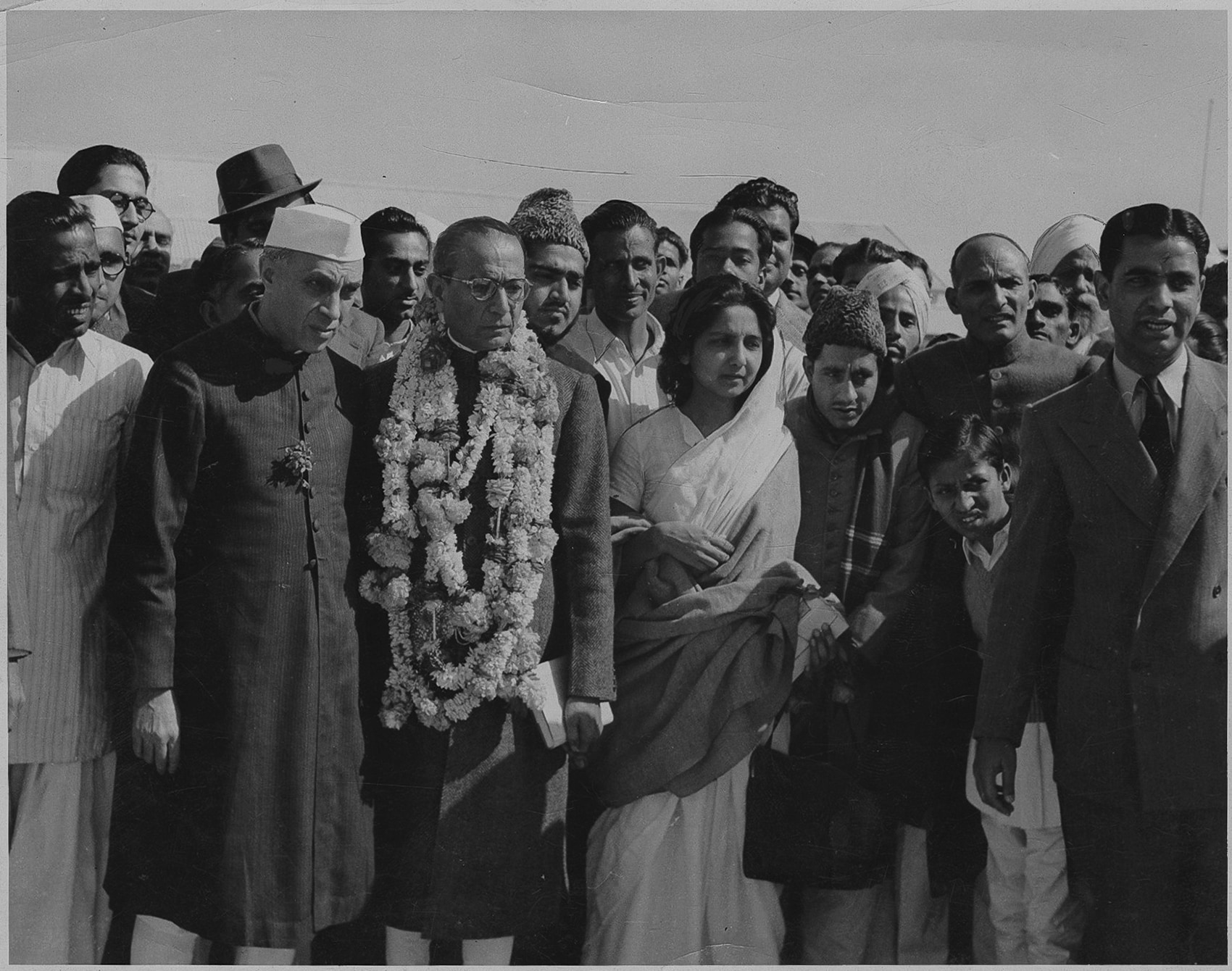 Black-and-white photograph of Asaf Ali wearing a heavy floral garland and round spectacles, standing beside Jawaharlal Nehru in a Gandhi cap, with Aruna Asaf Ali in a white sari to his left, amid a crowd at an outdoor gathering