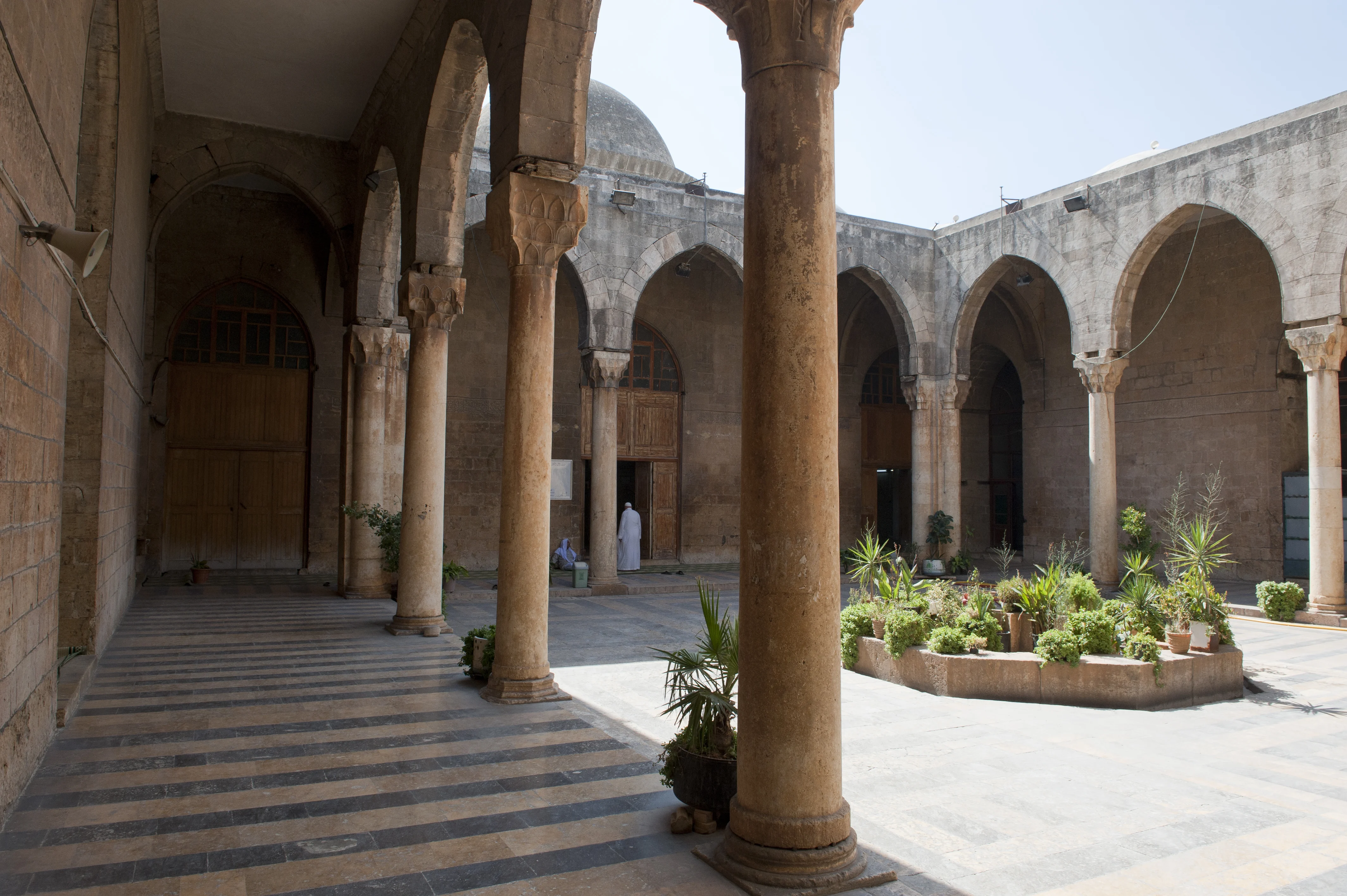 The stone facade of the Firdaws Madrasa in Aleppo, showing its carved portal with interlocking voussoirs and a decorated muqarnas hood above the entrance