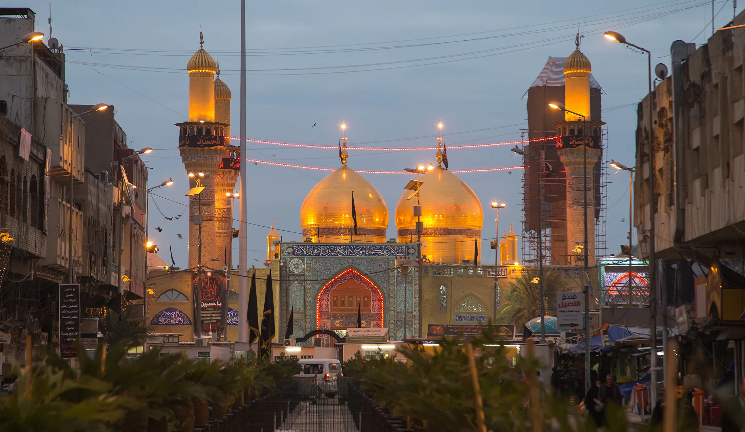 The golden-domed Al-Kazimiyya Mosque in Baghdad, with its twin gold minarets and dome complex reflected in the surrounding pool, seen across an open plaza