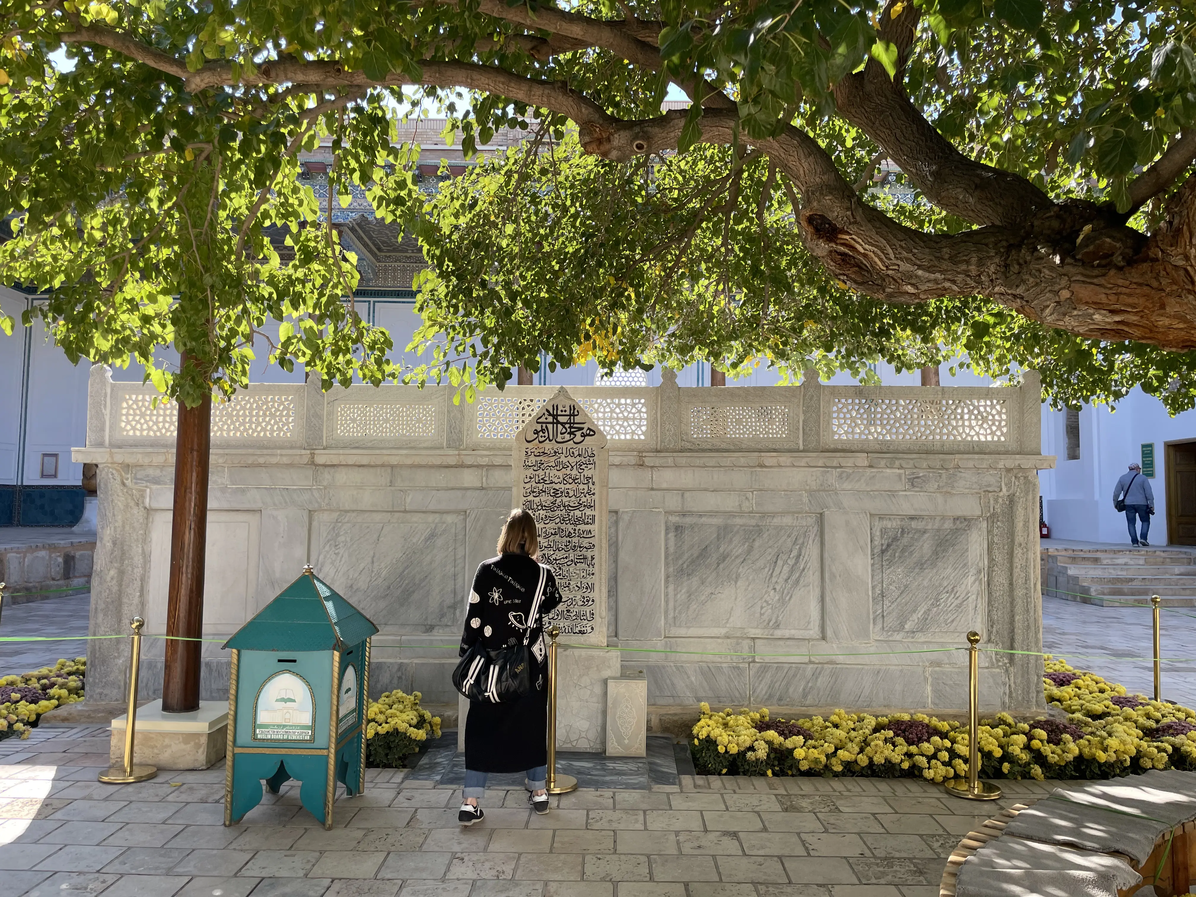 The grave enclosure of Baha al-Din Naqshband: a low white marble platform with pierced stone screens along its upper edge and a tall marble headstone carved with an inscription in black, shaded by the spreading branches of an old tree, with beds of yellow and purple chrysanthemums, brass posts carrying a rope barrier, and a turquoise donation box to the left; two visitors are present, one standing at the headstone and one walking away in the background