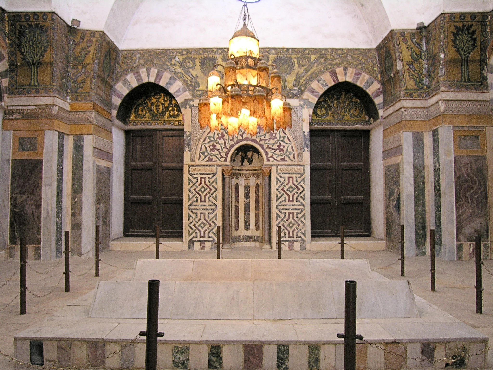 The interior of Baybars's mausoleum chamber: a low marble tomb platform behind chain posts, facing a marble-inlaid mihrab flanked by tall wooden doors, with gold mosaic tree panels above and a large hanging chandelier