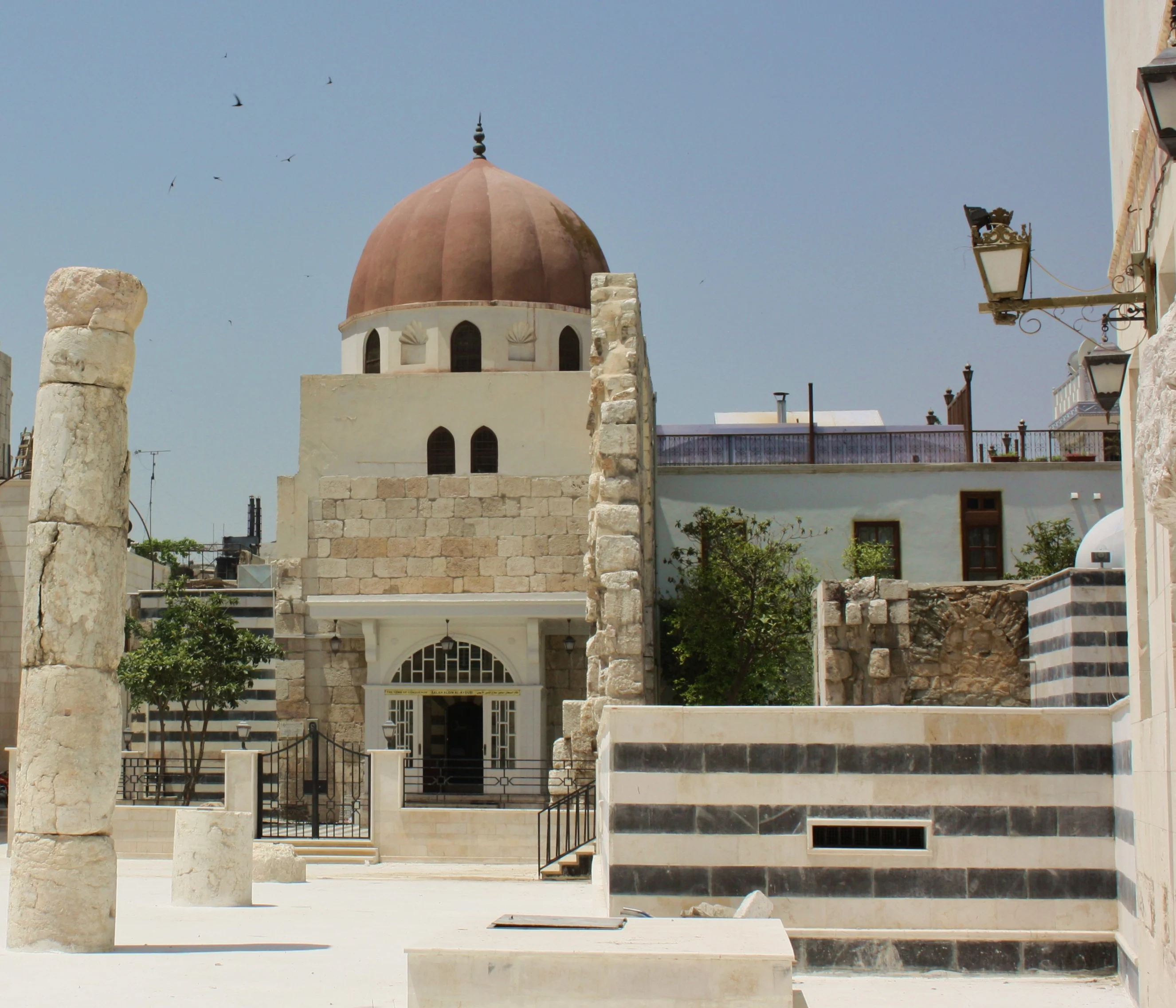 The mausoleum of Saladin, a stone building crowned by a ribbed red-brown dome, its arched entrance carrying a nameplate, with antique columns and striped masonry in the courtyard before it