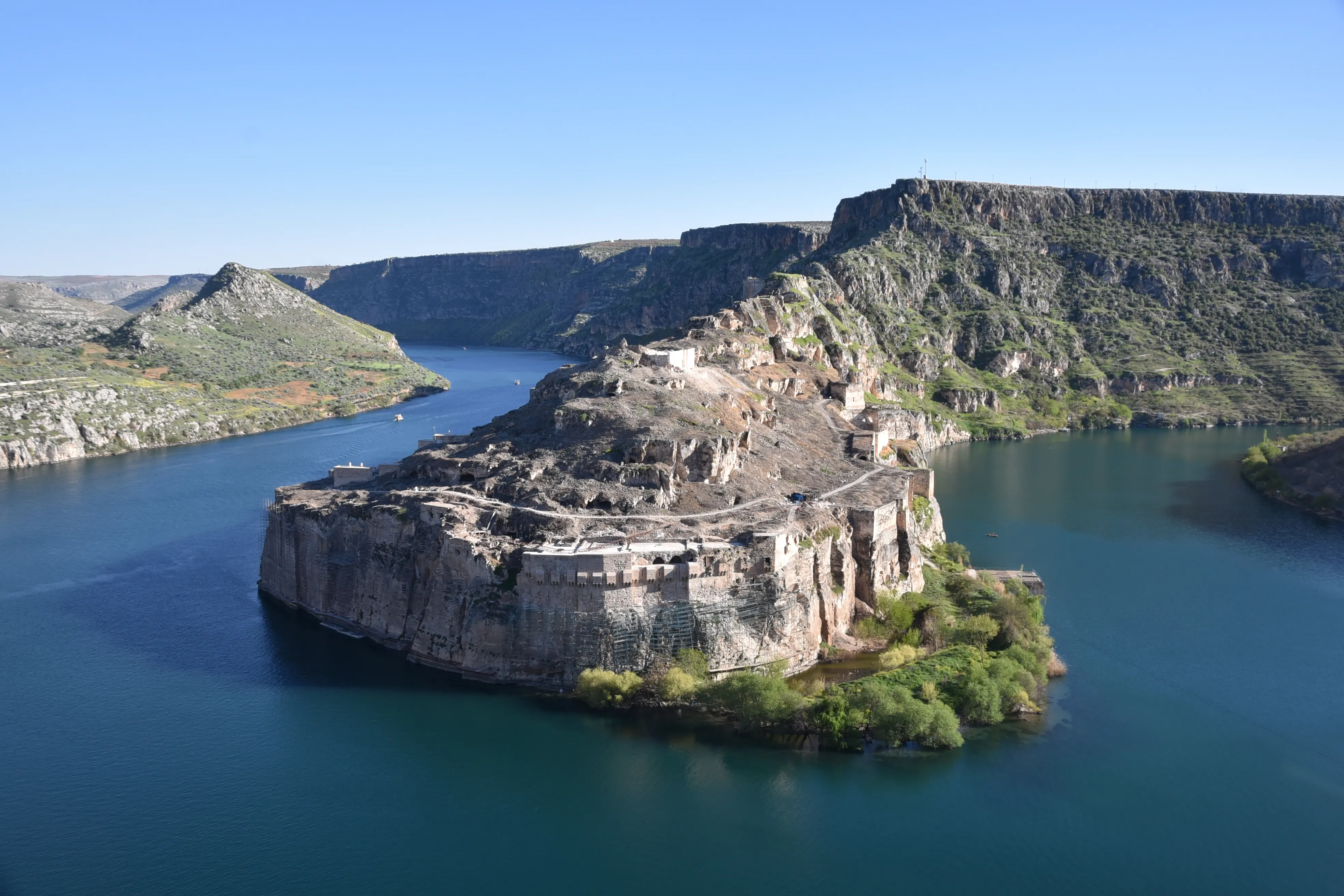 A fortified rocky promontory carrying ruined walls, rising above the wide blue water of the Euphrates and enclosed by cliffs and high plateau
