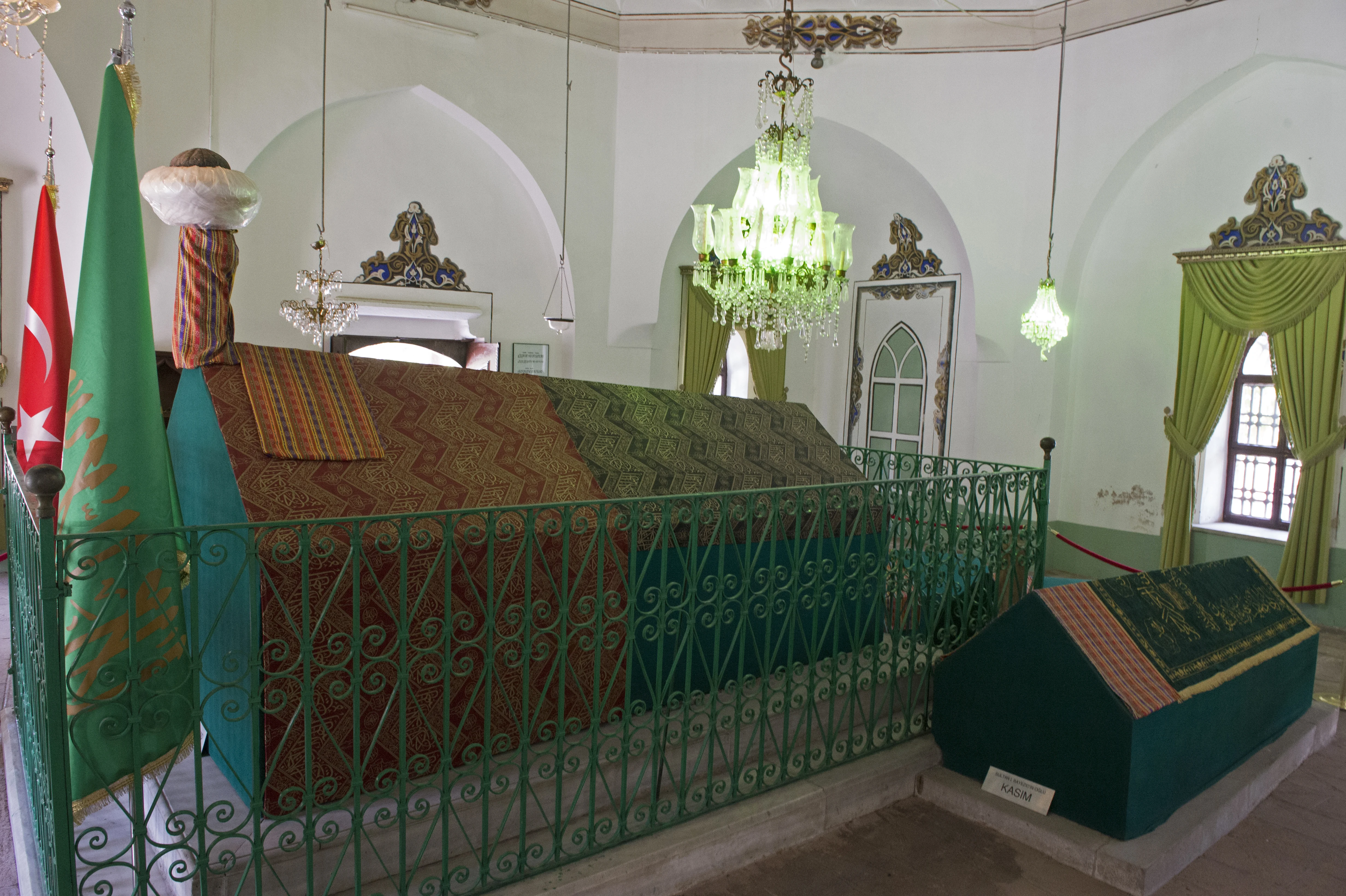 The cloth-draped cenotaph of Bayezid I inside his türbe, enclosed by a green iron railing beneath a chandelier, with a turban finial at its head, banners alongside and a smaller tomb to the right