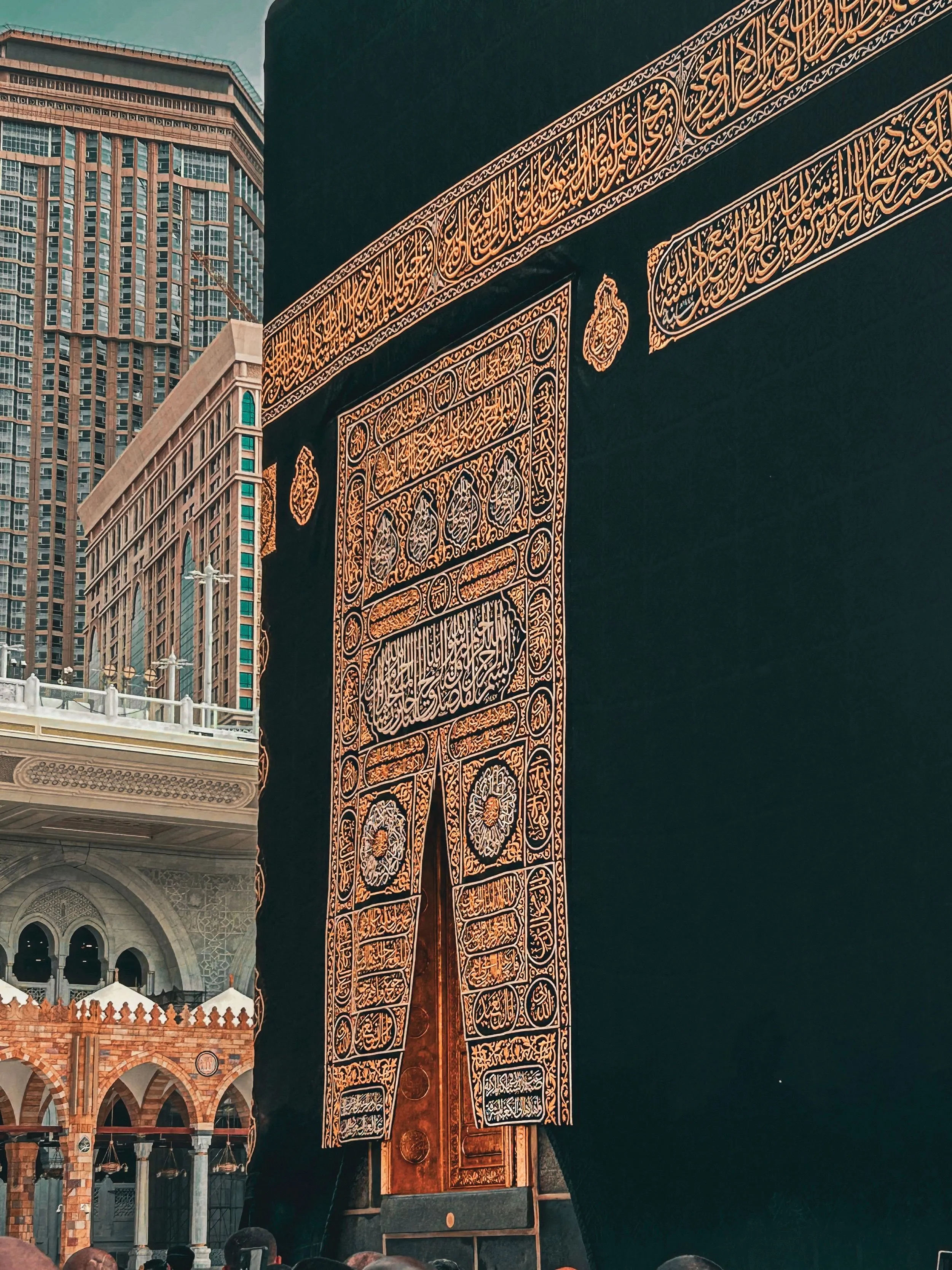 The gilded door of the Kaaba set in the black kiswah embroidered with gold calligraphy, with the arcades of the Masjid al-Haram and a tall modern tower rising behind