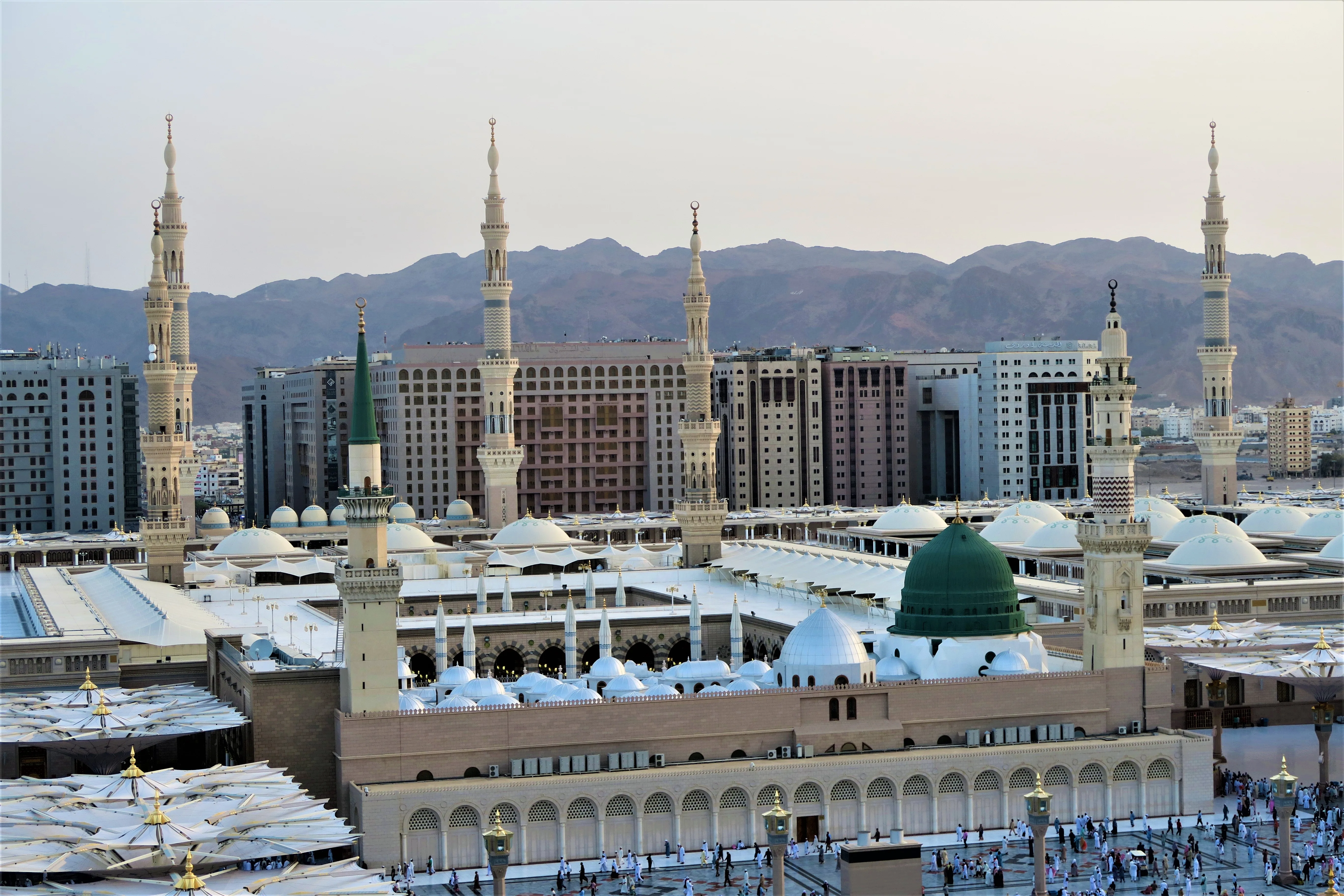An elevated view of Al-Masjid an-Nabawi in Medina, showing the green dome, tall minarets, white courtyard canopies and the surrounding city with mountains beyond