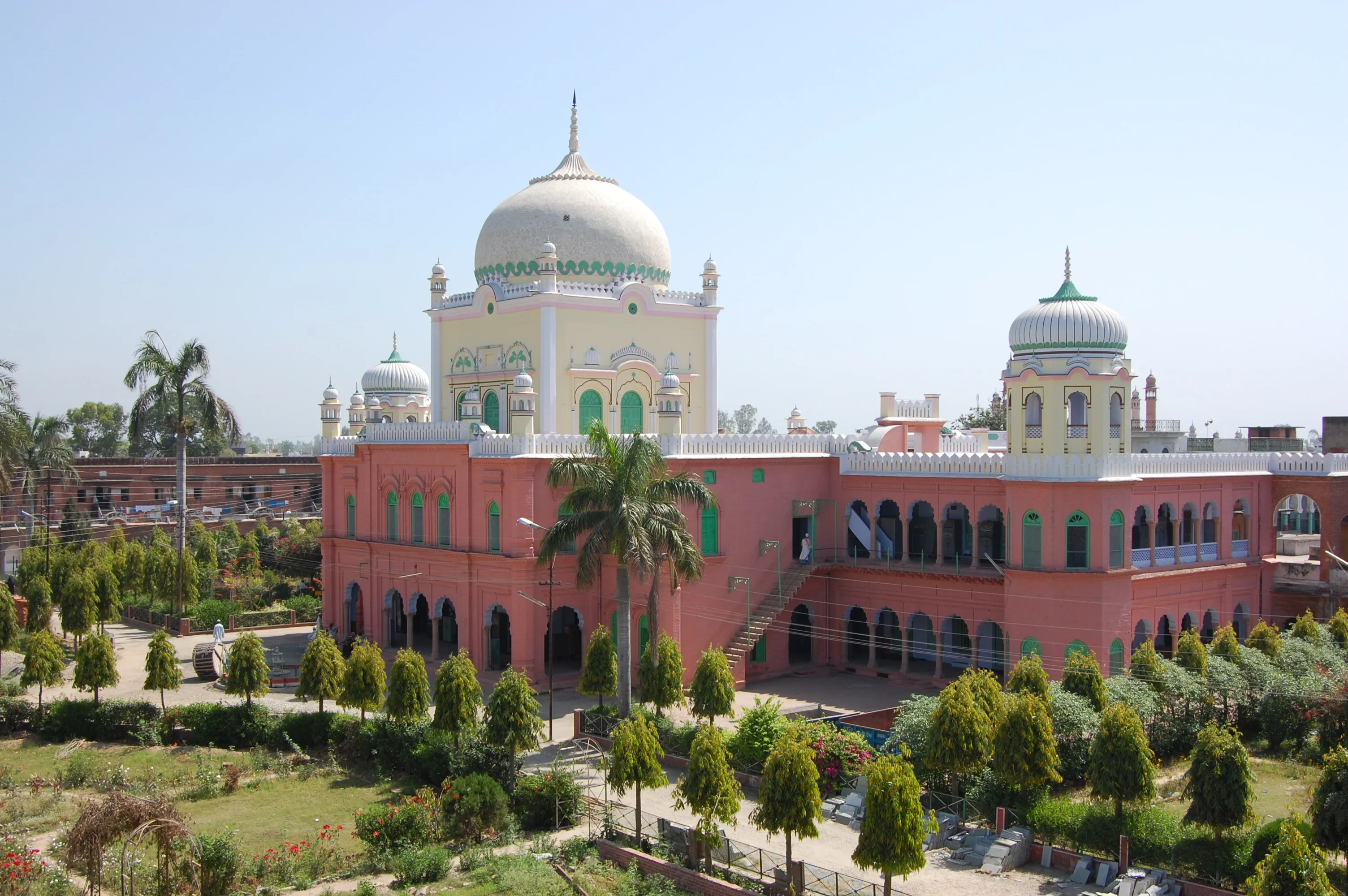 A pink two-storey arcaded seminary building with rows of pointed-arch openings, with a cream-coloured white-domed mosque behind it and gardens and palm trees in the foreground