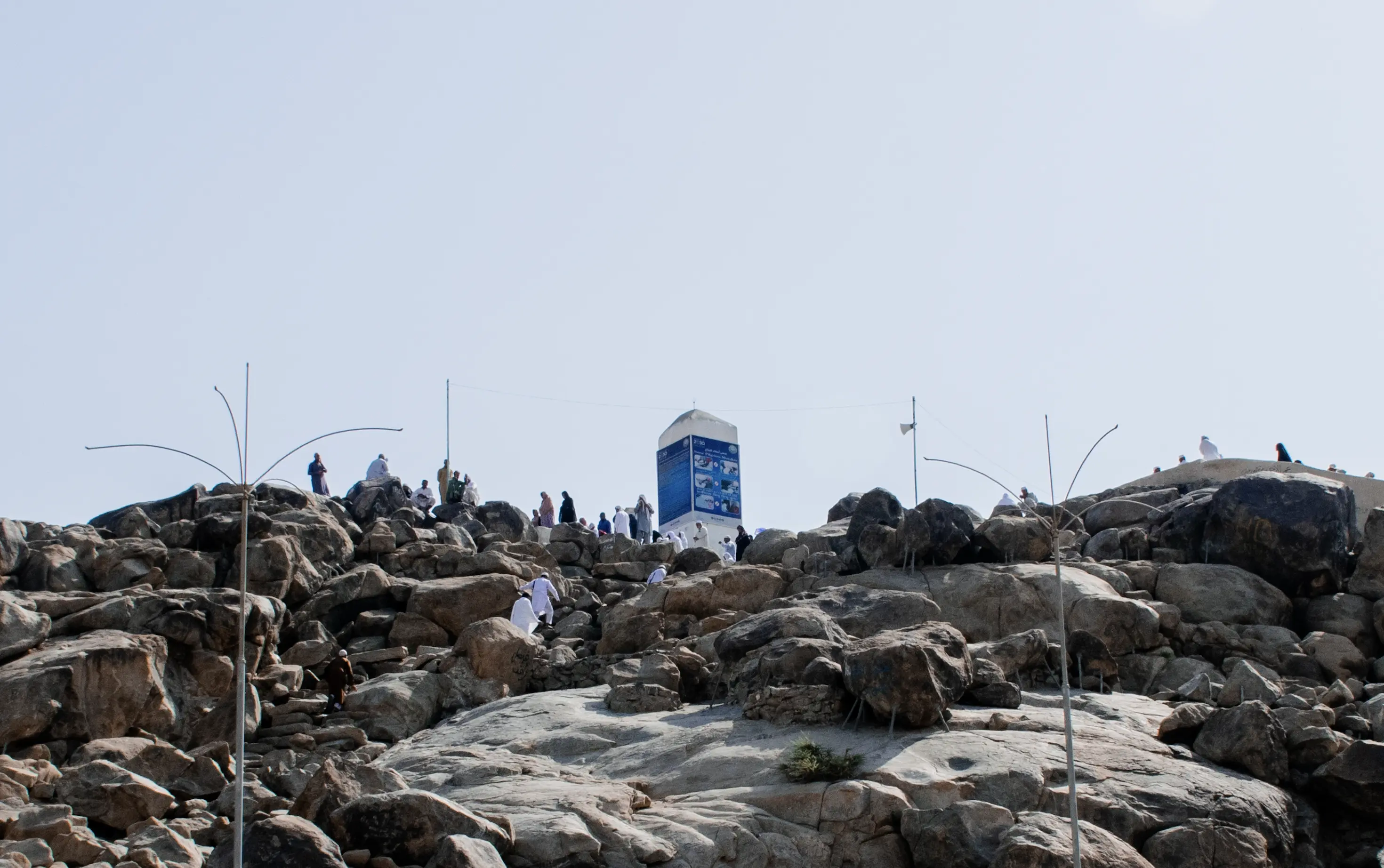 The boulder-strewn granite summit of a low hill under a pale sky, a white tapering pillar standing at the top with an illustrated information panel fixed to it, slender lamp and lightning poles rising among the rocks with a cable strung between them and a loudspeaker on one, and pilgrims in white and in darker dress climbing over the boulders and gathered around the pillar