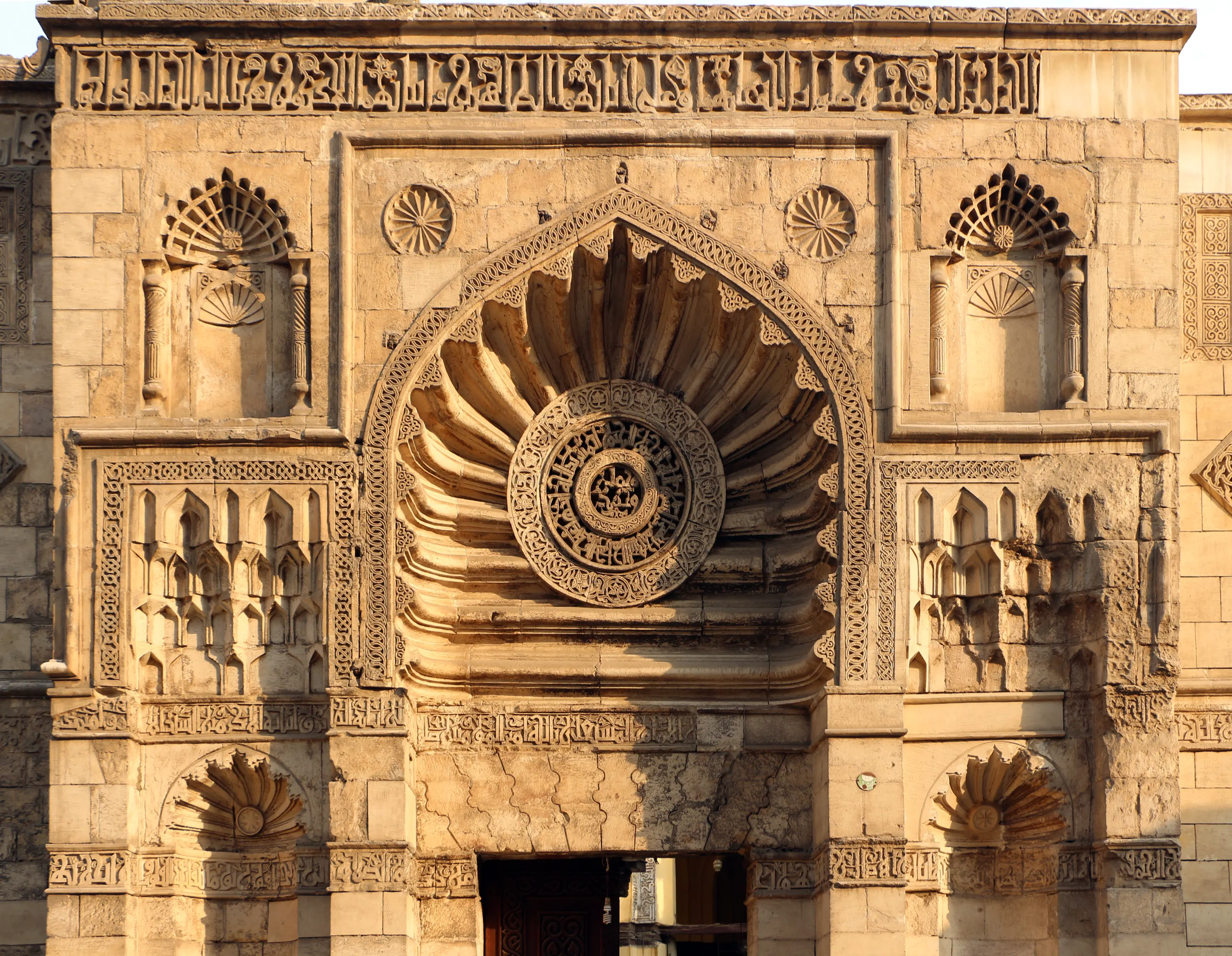A close view of the carved stone entrance facade of the Al-Aqmar Mosque, with a shell-shaped hood over a recessed niche framed in decorative strapwork, flanked by smaller fan-shaped niches and bands of Kufic inscription