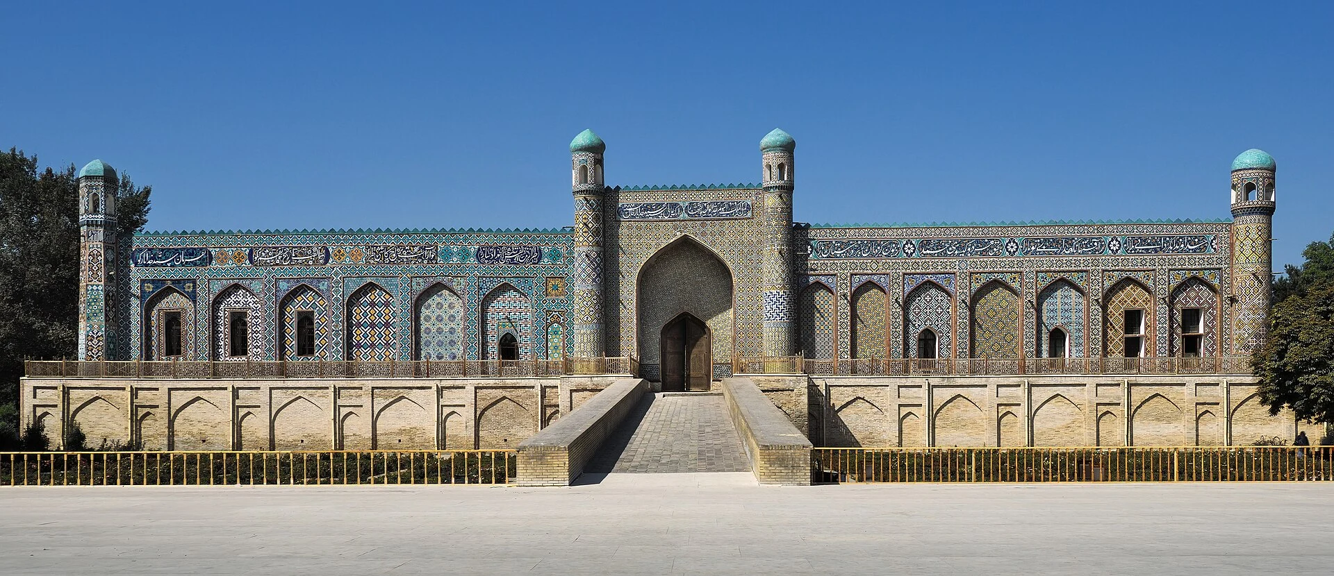 The long tiled facade of the Khan's Palace at Kokand, with a high arched central portal, turquoise-domed corner minarets and bands of polychrome tilework and Arabic inscription