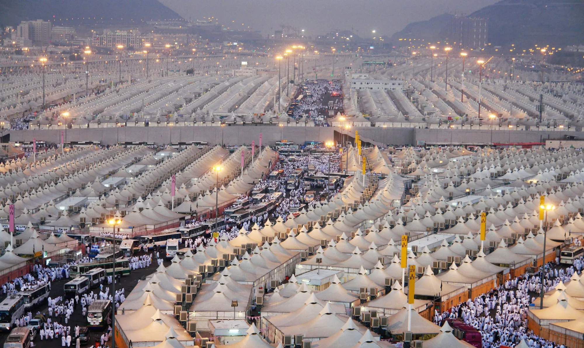 An elevated view at dusk over the tent city at Mina, rank upon rank of white peaked tents with air-conditioning units between them, streetlights and coaches along the roadways that divide the blocks, pilgrims in white moving between them, and the hills and buildings of the valley beyond