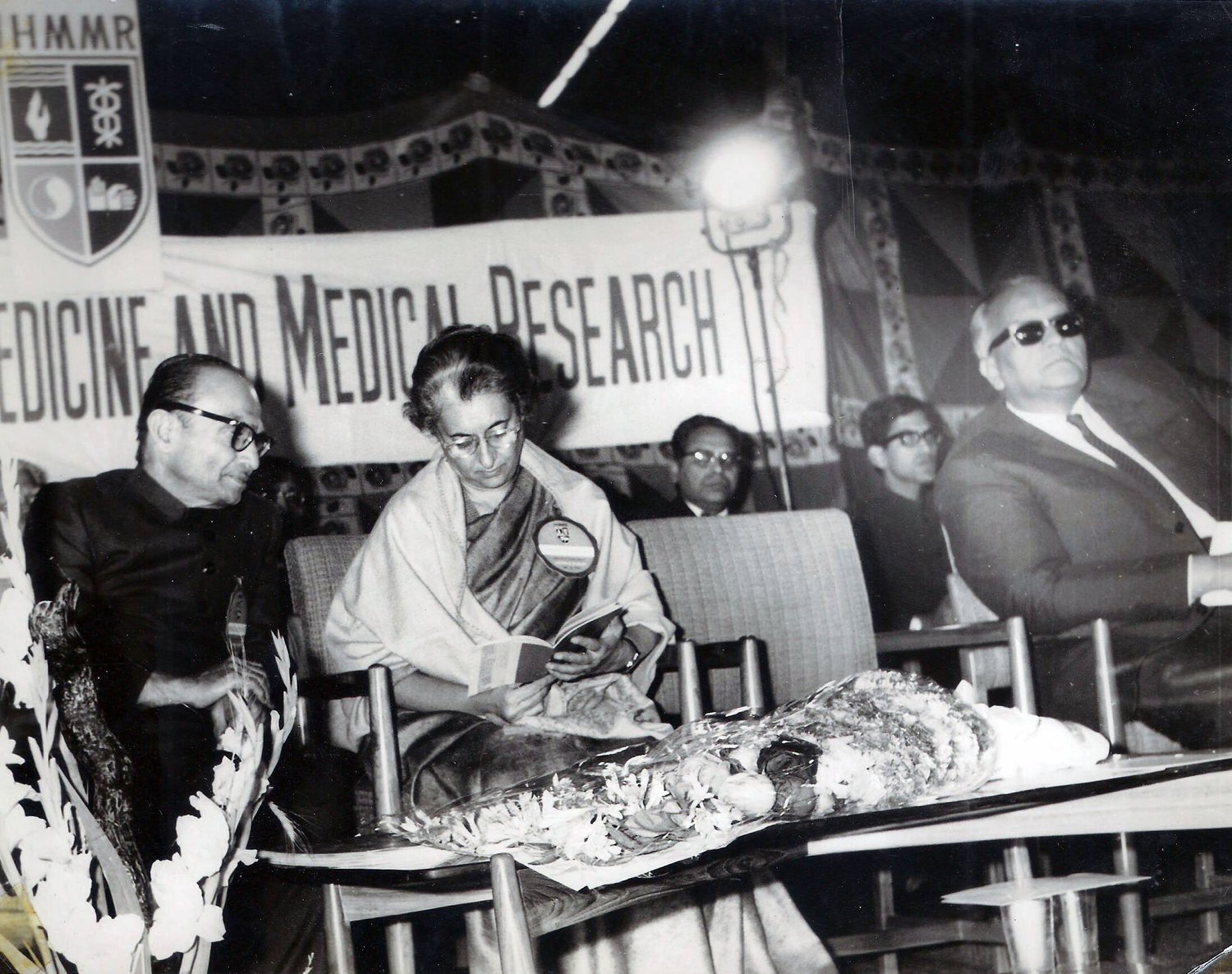 Black-and-white photograph of Hakeem Abdul Hameed, wearing spectacles and a dark jacket, seated on the left beside Indira Gandhi, who is seated at the centre reading a book, at a medicine and medical research event with other men behind them