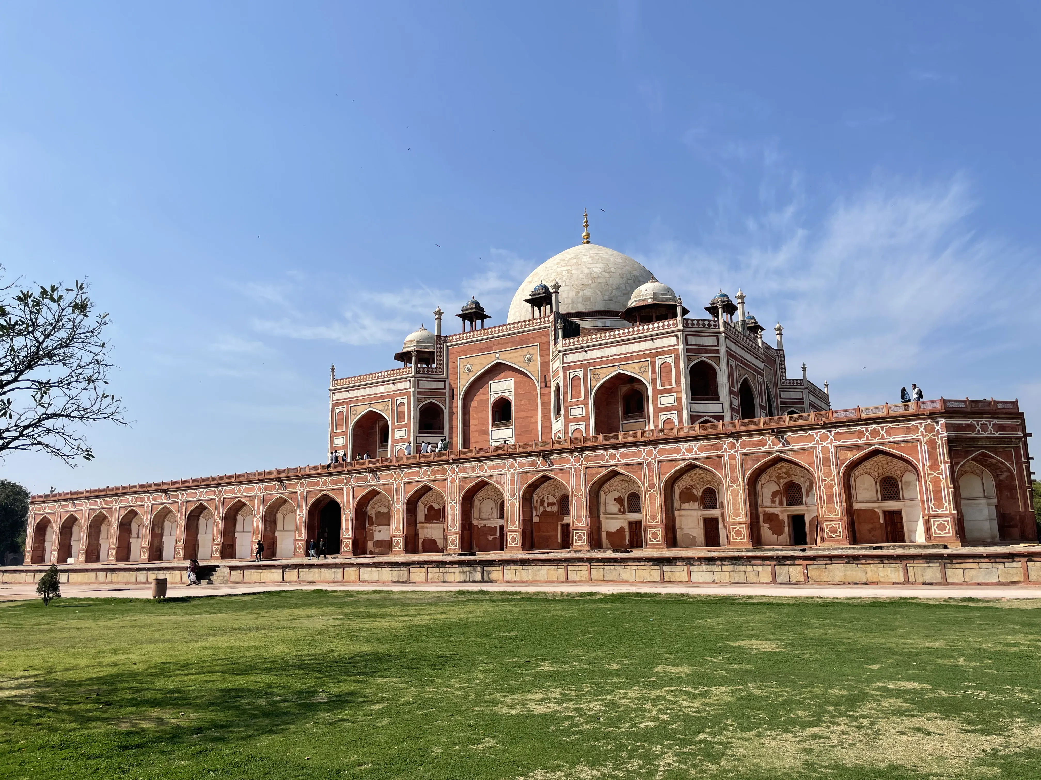 Humayun's Tomb in Delhi seen from the lawn: a red sandstone and white marble mausoleum with a high central dome flanked by domed corner pavilions, its facade patterned in white marble geometric inlay and pierced by tall arched recesses, set on an arcaded plinth