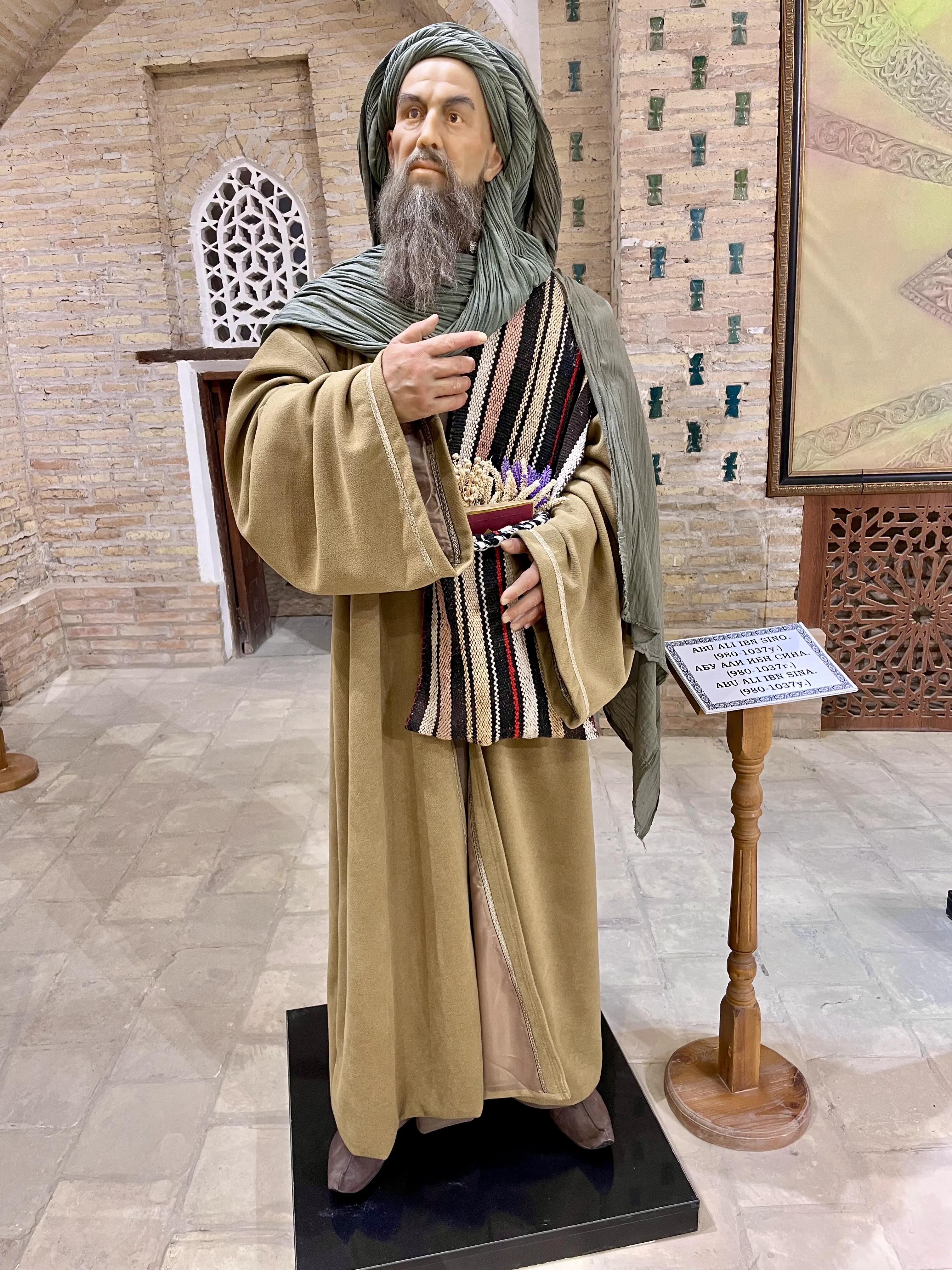 A life-size costumed figure of a bearded scholar in a sage-green turban and shawl over an olive robe, one hand raised to the chest, a woven striped sash at the waist holding a box of dried herbs and lavender, standing on a low plinth in a brick-vaulted hall beside a wooden lectern carrying a name label in three scripts
