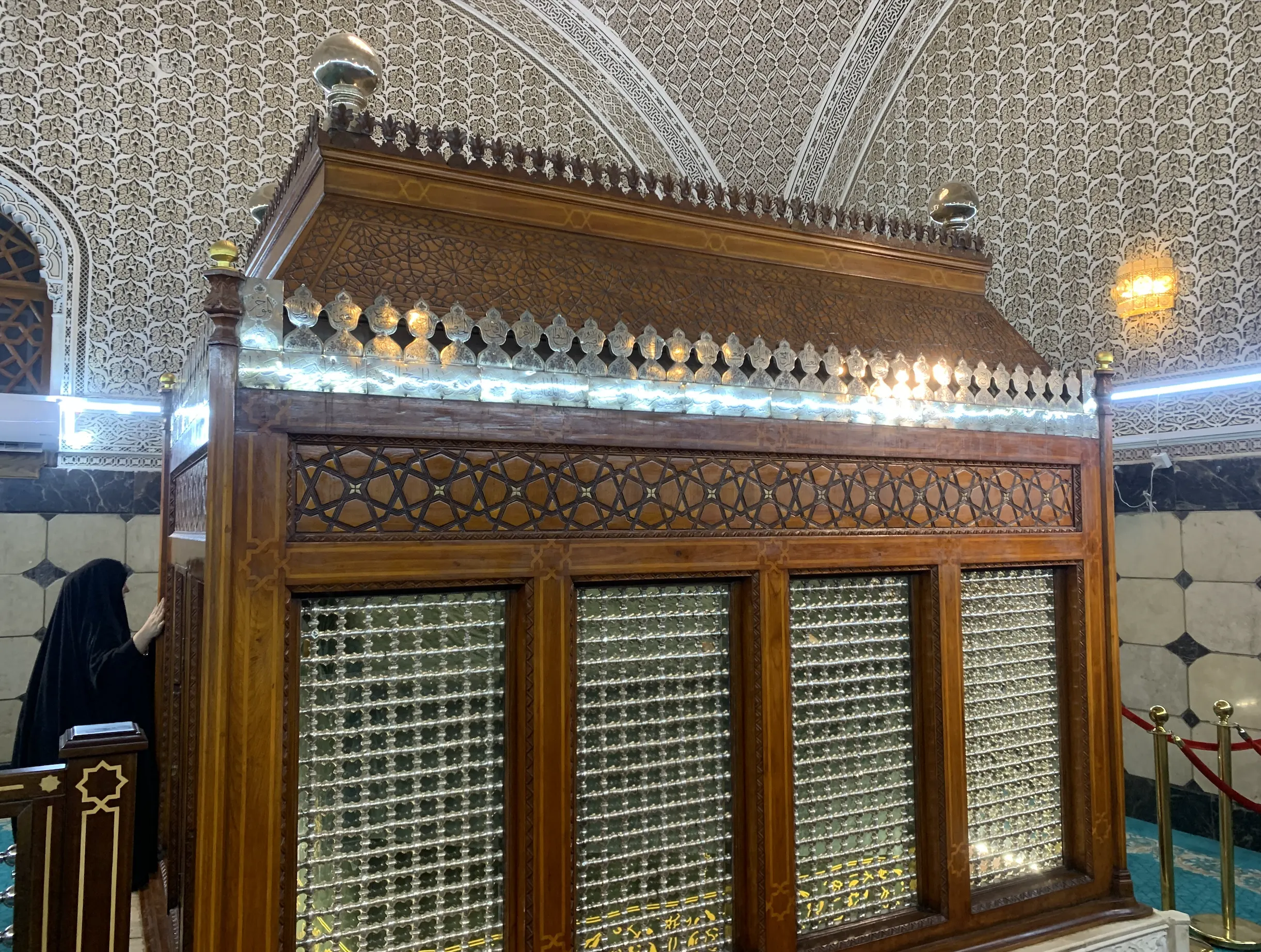 A tall carved teak enclosure over a grave, its panels worked in geometric latticework and fitted with glazed screens, topped by a rail of silver cresting bearing Arabic inscriptions and domed silver finials, standing on a white marble base within a chamber whose walls and vaults are covered in fine white arabesque tracery; a visitor in a black cloak stands at the near corner with one hand resting on the woodwork