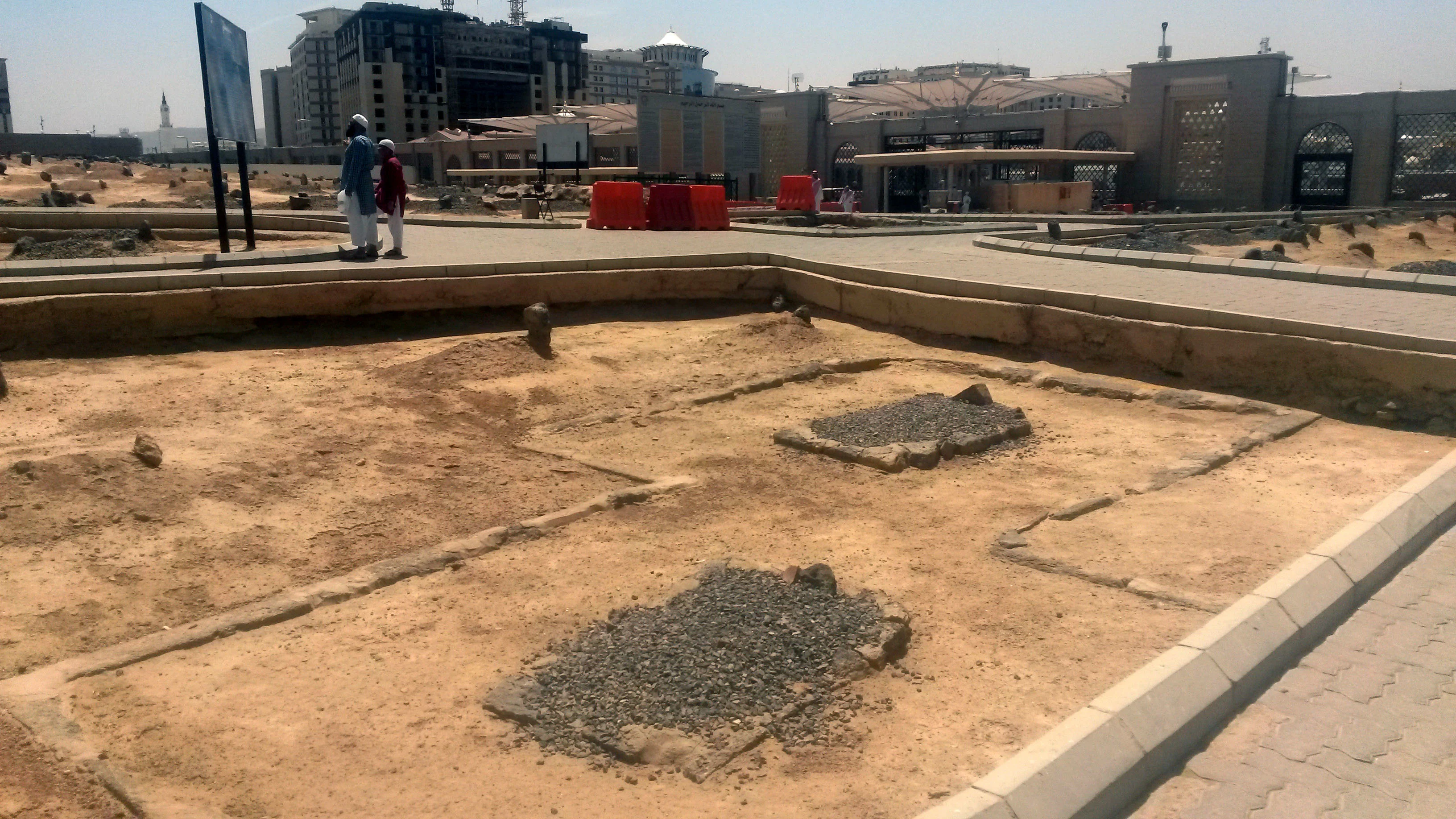 An open expanse of bare sandy ground at the Al-Baqi cemetery in Medina, divided into rectangular plots by low stone kerbs, with unmarked graves covered in loose grey gravel and small rough stones set at their heads, an information board to the left, paved walkways crossing the ground, and the boundary buildings of the cemetery and the modern towers of Medina rising behind