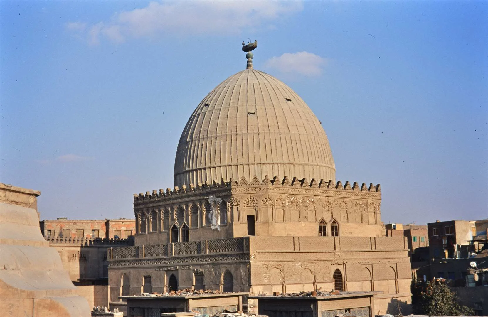 The mausoleum of Imam al-Shafi'i in Cairo seen from the rooftops: a large ribbed stone dome carrying a metal finial with a boat-shaped ornament at its top, rising from a square block edged with stepped crenellations, a band of small paired arched niches and carved panels below the dome, and pointed-arched grilled windows in the storey beneath, with the flat roofs of the surrounding city around it