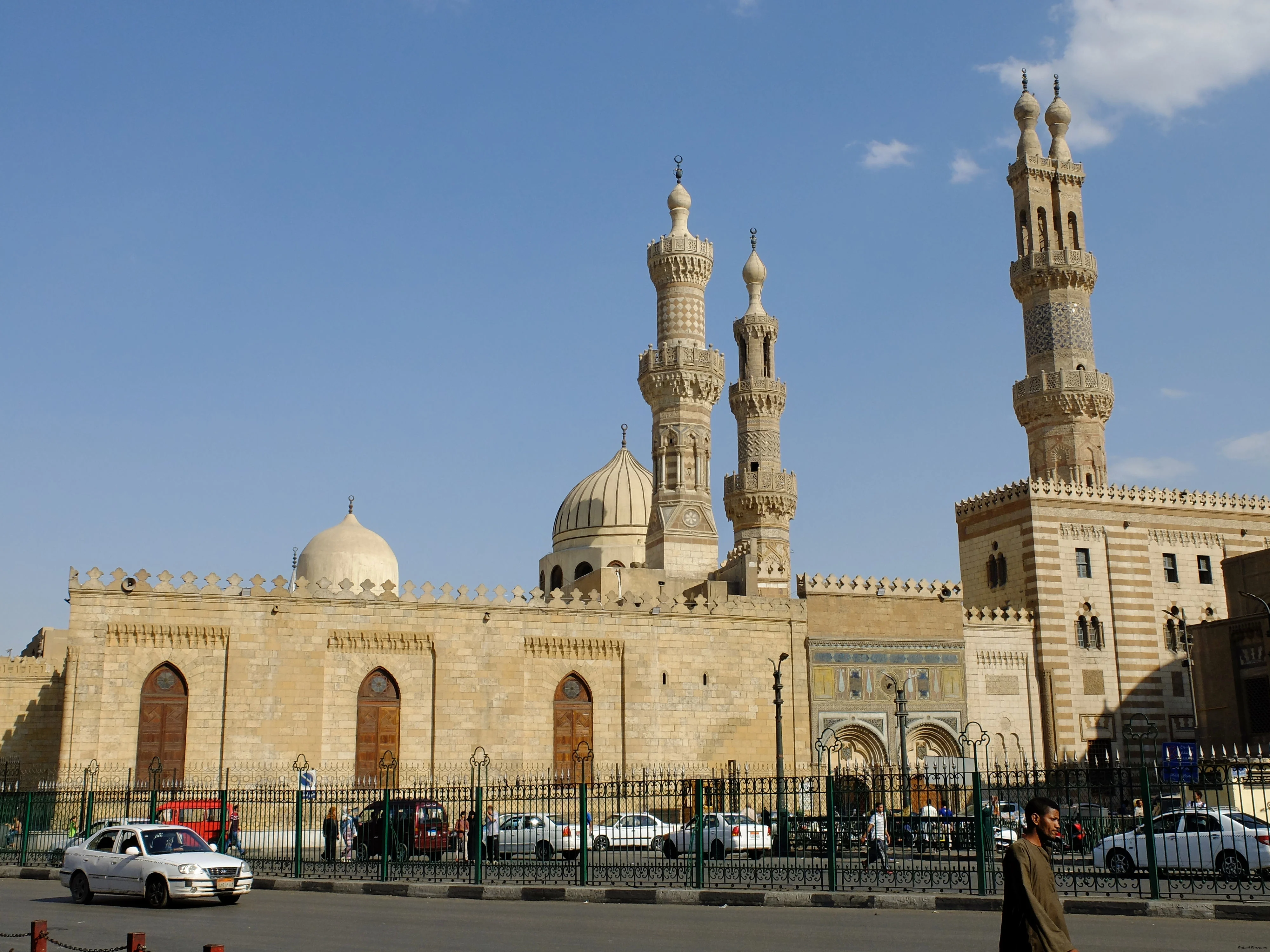The stone facade of Al-Azhar Mosque behind railings on a Cairo street, with two pairs of minarets of differing styles rising above the courtyard wall, one pair topped with onion domes, and pedestrians and cars passing in front