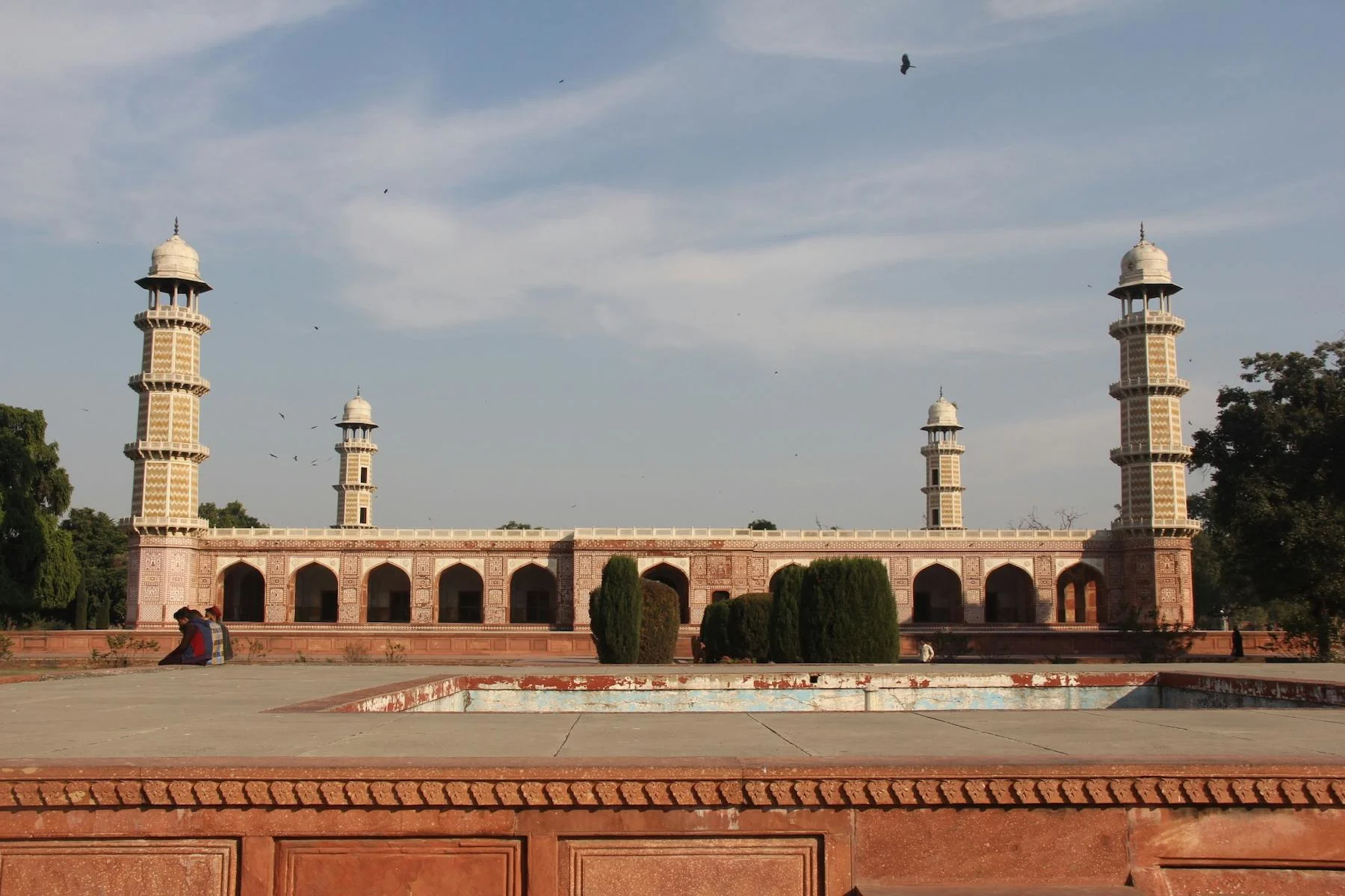 The tomb of Jahangir at Shahdara, Lahore: a long single-storey red sandstone and white marble building with four tall tapering minarets banded in cream and ochre marquetry, one at each corner, seen across an empty reflecting pool