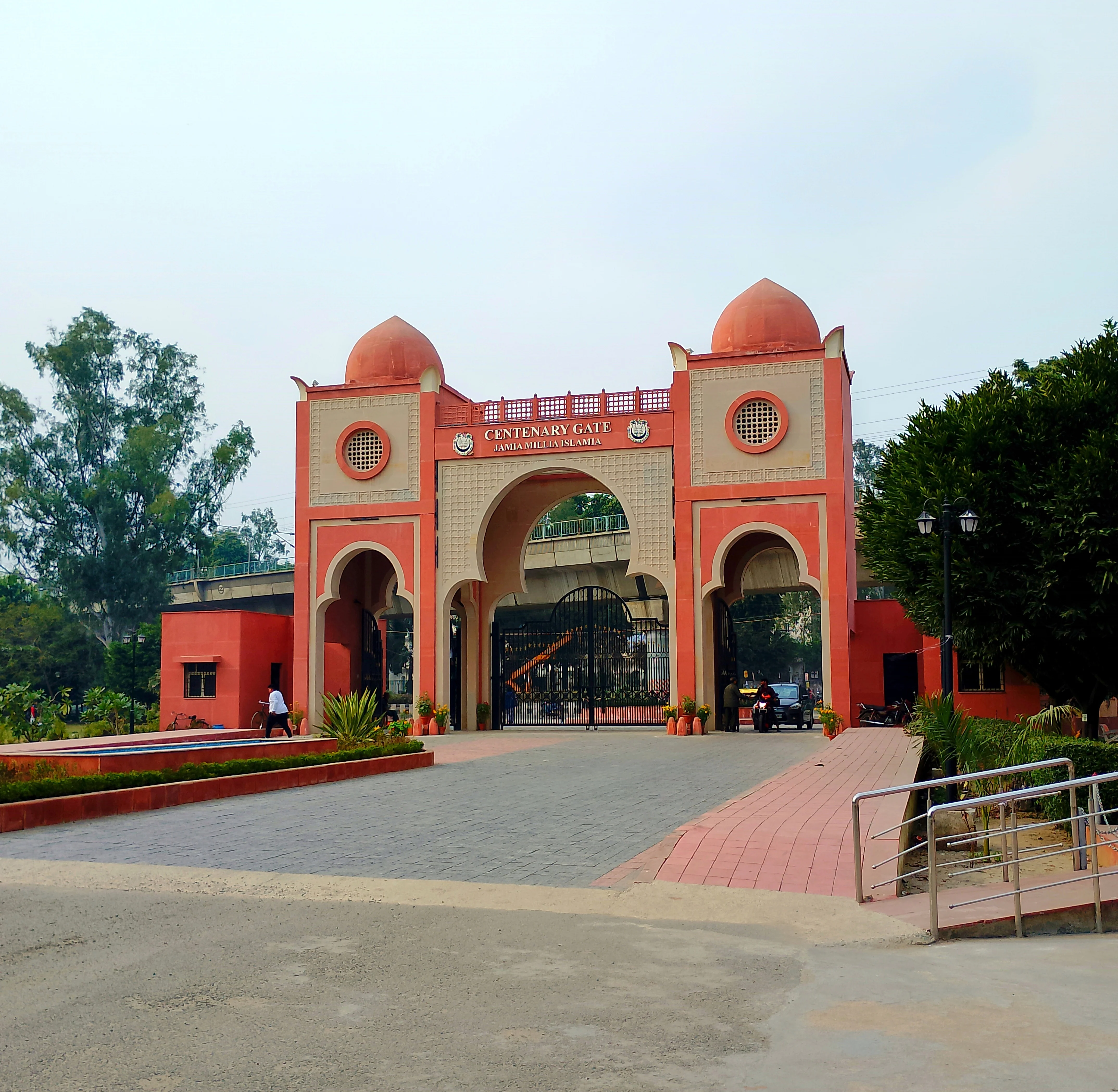 A monumental red-and-cream gateway inscribed 'Centenary Gate Jamia Millia Islamia', with a raised metro flyover passing behind it