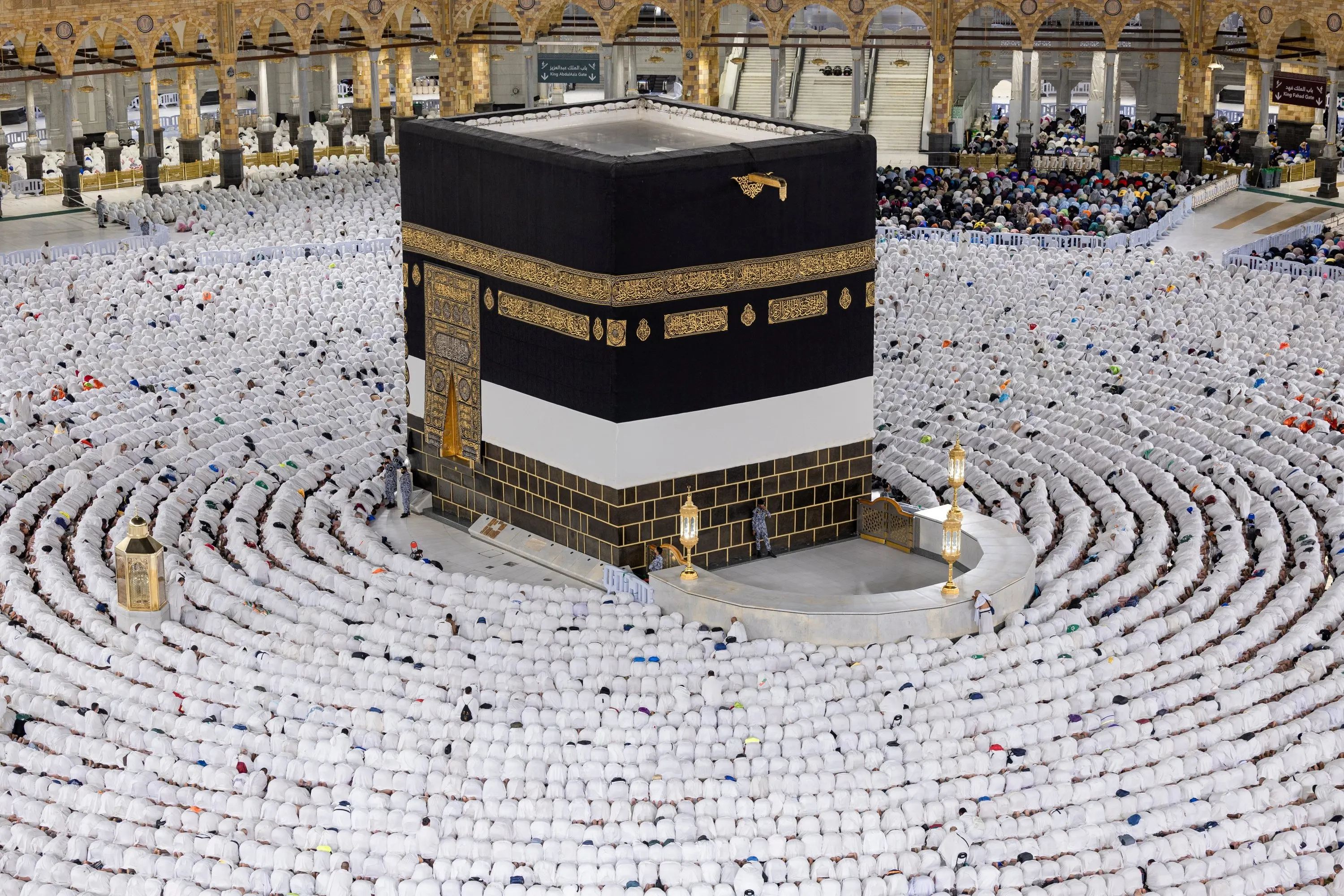 The Kaaba draped in the black kiswah, its upper wall carrying a band of gold calligraphy and its gold-embroidered door set above a marble base course, standing at the centre of the Masjid al-Haram; concentric rows of worshippers in white ihram fill the courtyard around it, with gilded lanterns and the curved enclosure beside the Kaaba, and the arcaded galleries of the mosque and its signed gateways behind