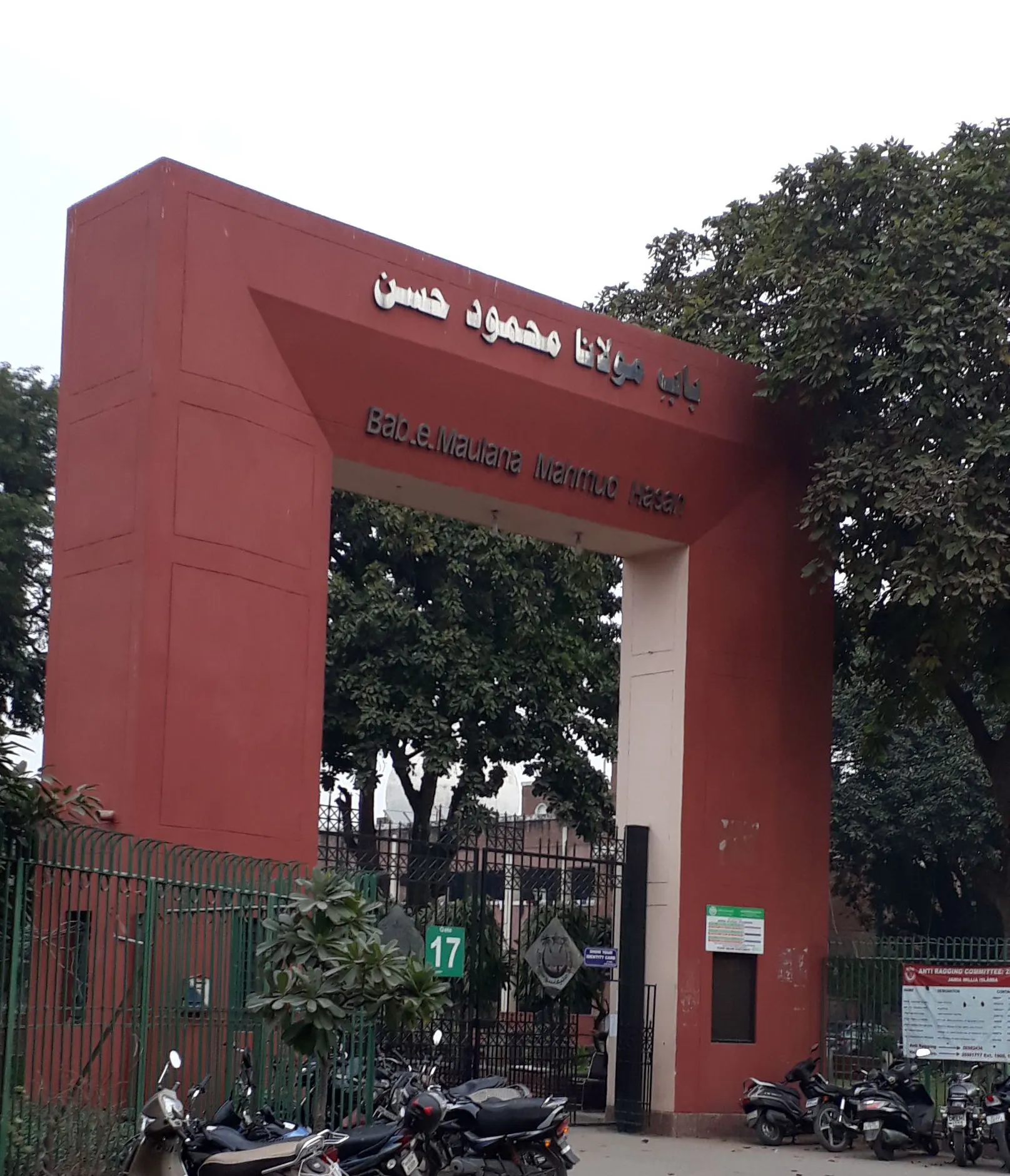 A tall red-painted stone gateway lettered in Urdu script and in Roman letters reading Bab-e-Maulana Mahmud Hasan, standing behind a green metal railing, with trees, a notice board and parked motorcycles in front of it