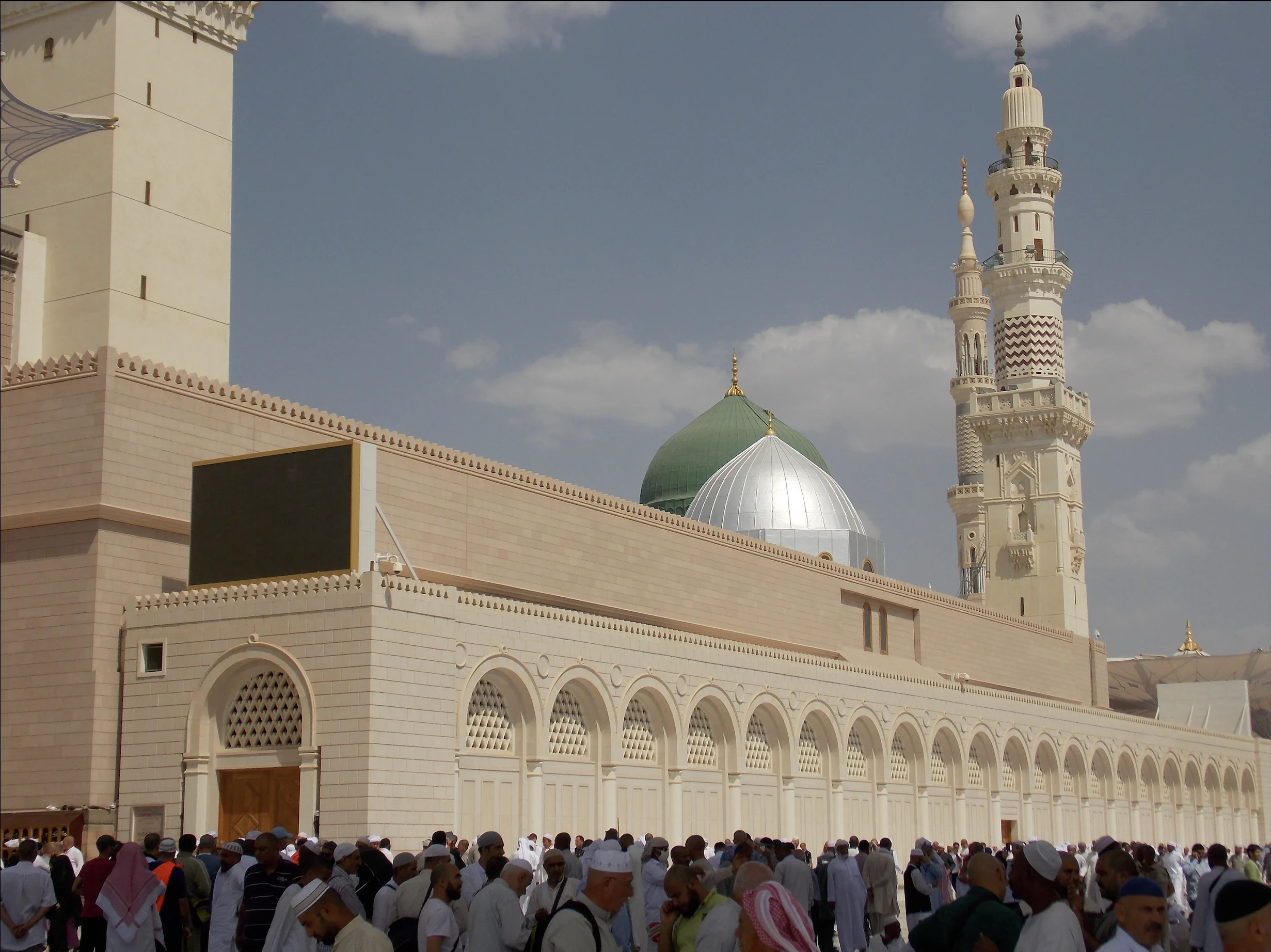The green dome of Al-Masjid al-Nabawi rising behind the pale stone wall of the mosque, with a silver dome in front of it and a tall balconied minaret to the right; a long arcade of arched recessed panels runs along the wall below, a large display screen is mounted on the stonework, and a crowd of worshippers fills the plaza in the foreground