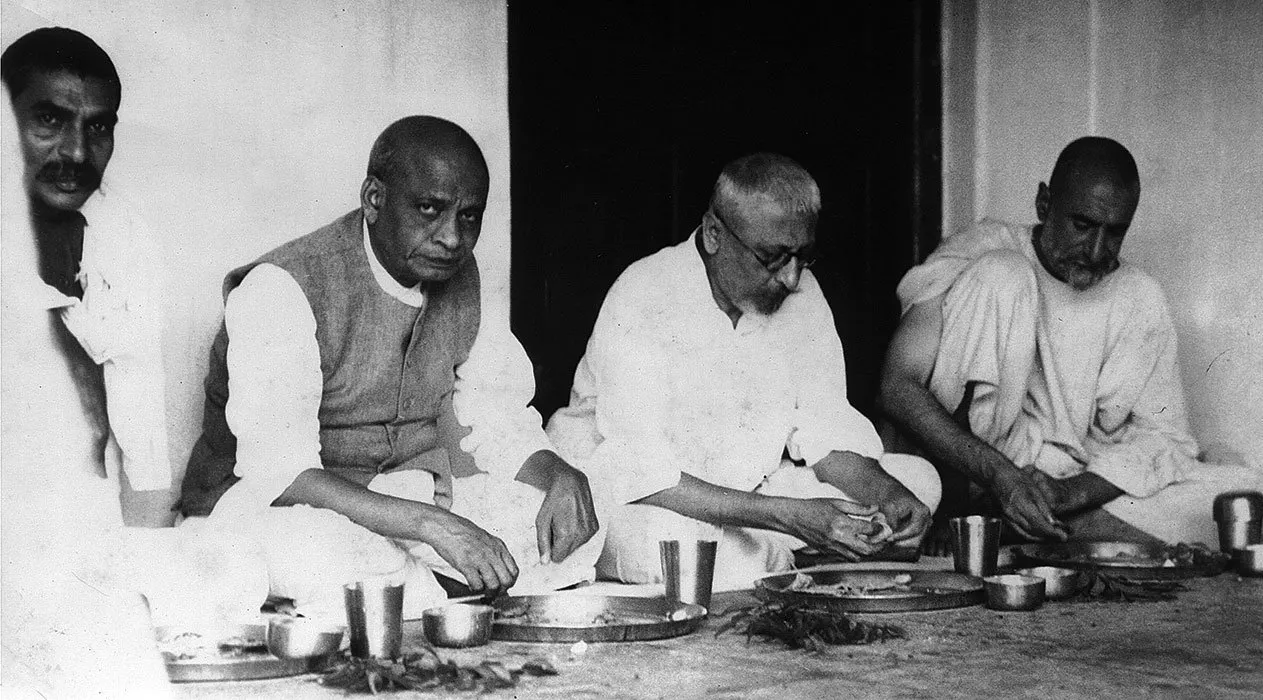 Black-and-white photograph of four men seated on the floor in a row, eating from metal platters with small bowls and tumblers set before them, a dark doorway behind; the second from the left wears a sleeveless jacket over a white kurta, the third wears round spectacles, and the man on the right has a beard and a shawl drawn over his shoulder