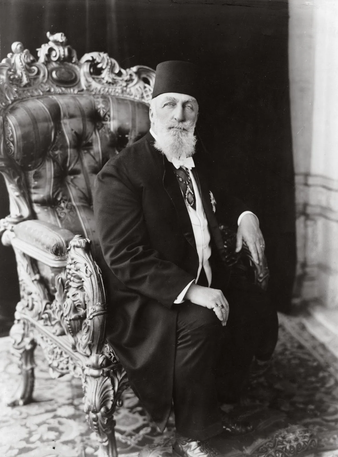 A formal seated portrait of an elderly man with a full white beard, wearing a fez, a dark frock coat with a sash and decorations, seated in an ornately carved armchair