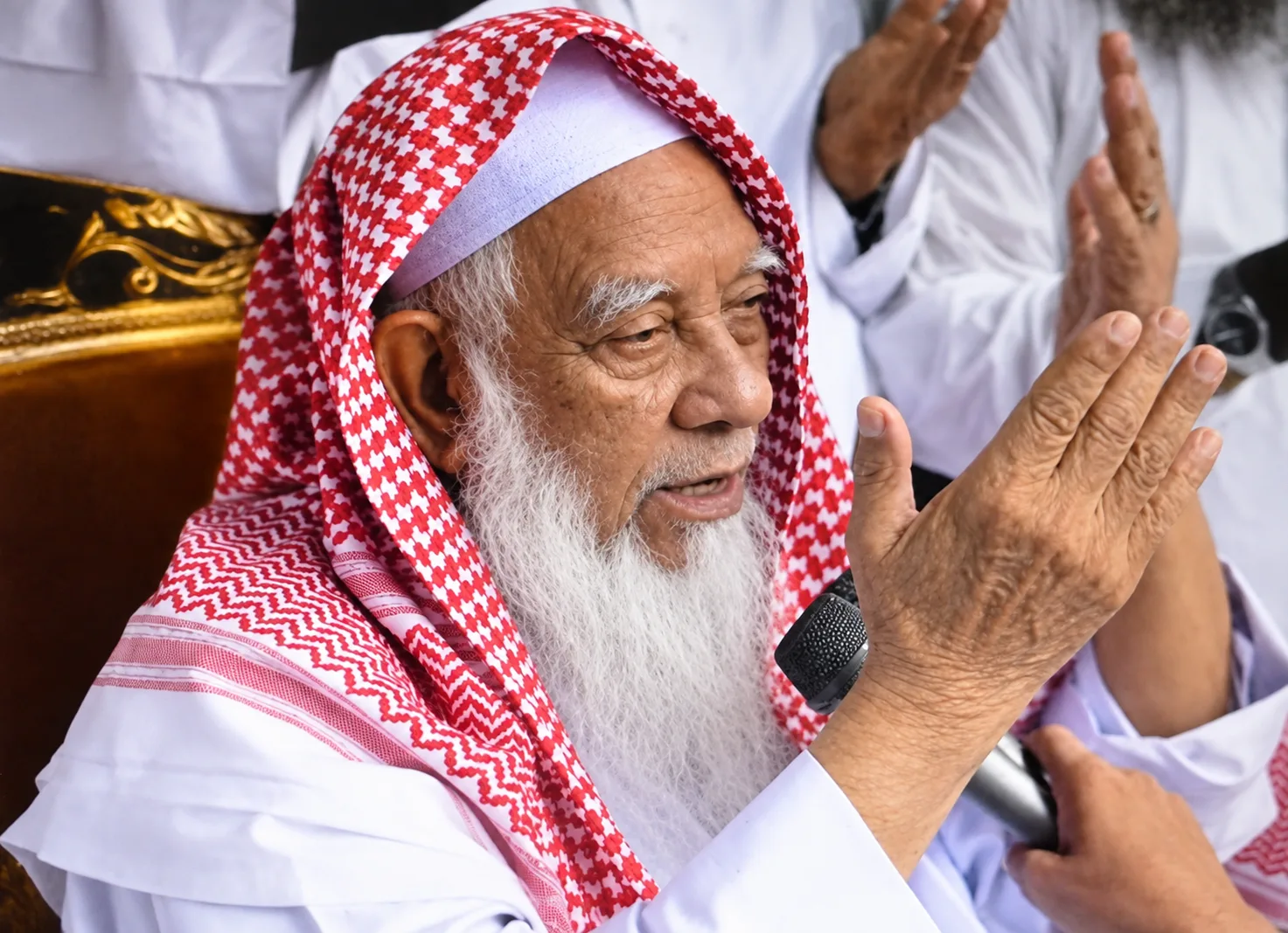 A very elderly man with a long white beard, wearing a white cap beneath a red and white checked headcloth, holding a microphone with his hands raised in supplication, with others behind him also raising their hands