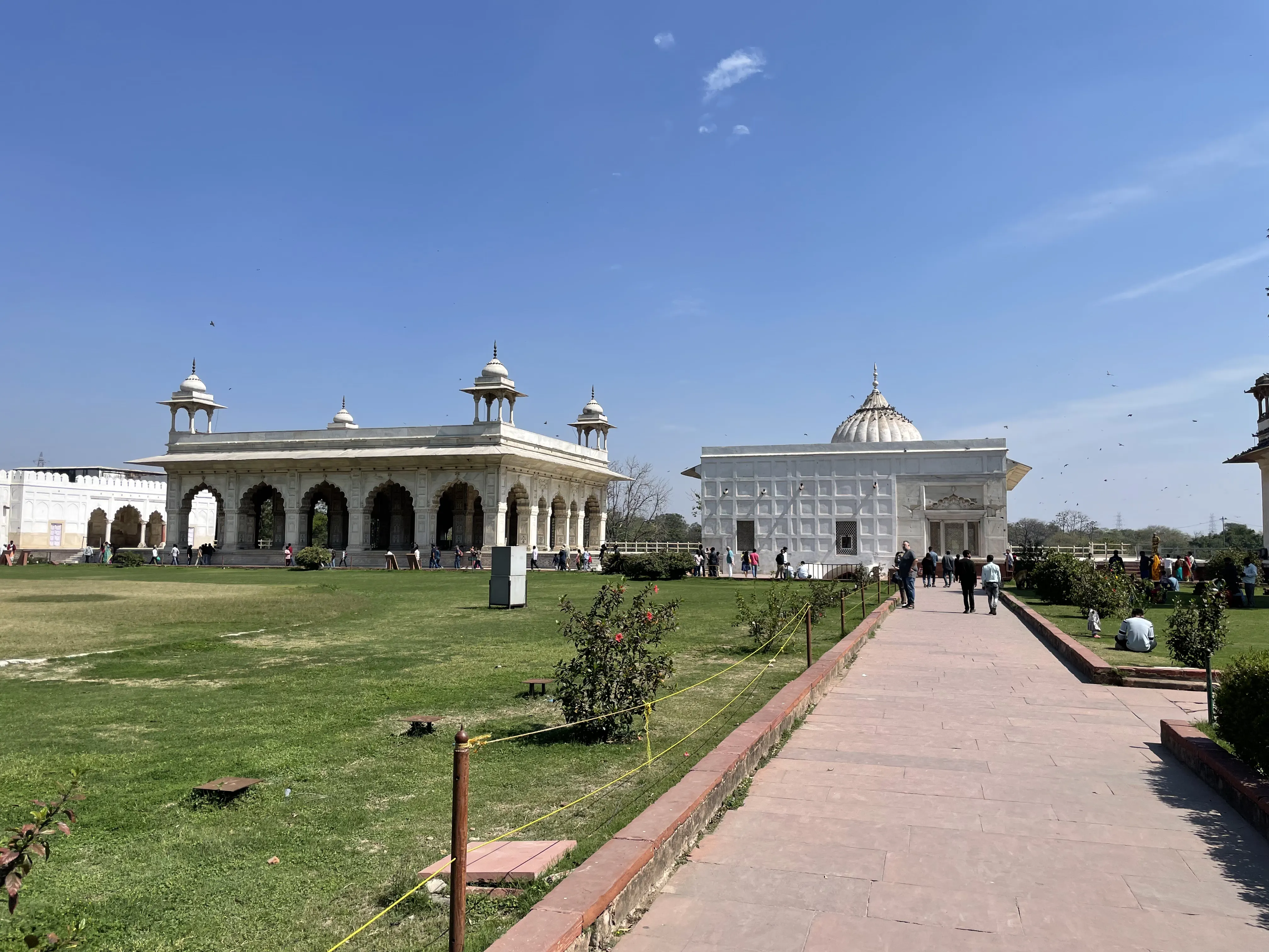 A courtyard within the Red Fort in Delhi: a white marble pavilion with cusped arches and domed corner chattris to the left, and a domed marble building faced with latticed stone screens to the right, set behind a lawn and a paved walkway with visitors