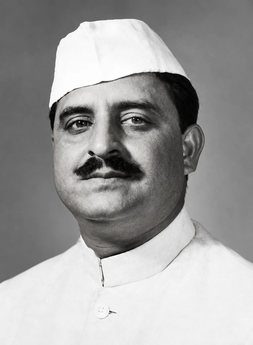 Black-and-white studio portrait of a man, head and shoulders, with a full dark moustache, wearing a white cap and a white closed-collar coat buttoned at the throat, against a plain grey backdrop
