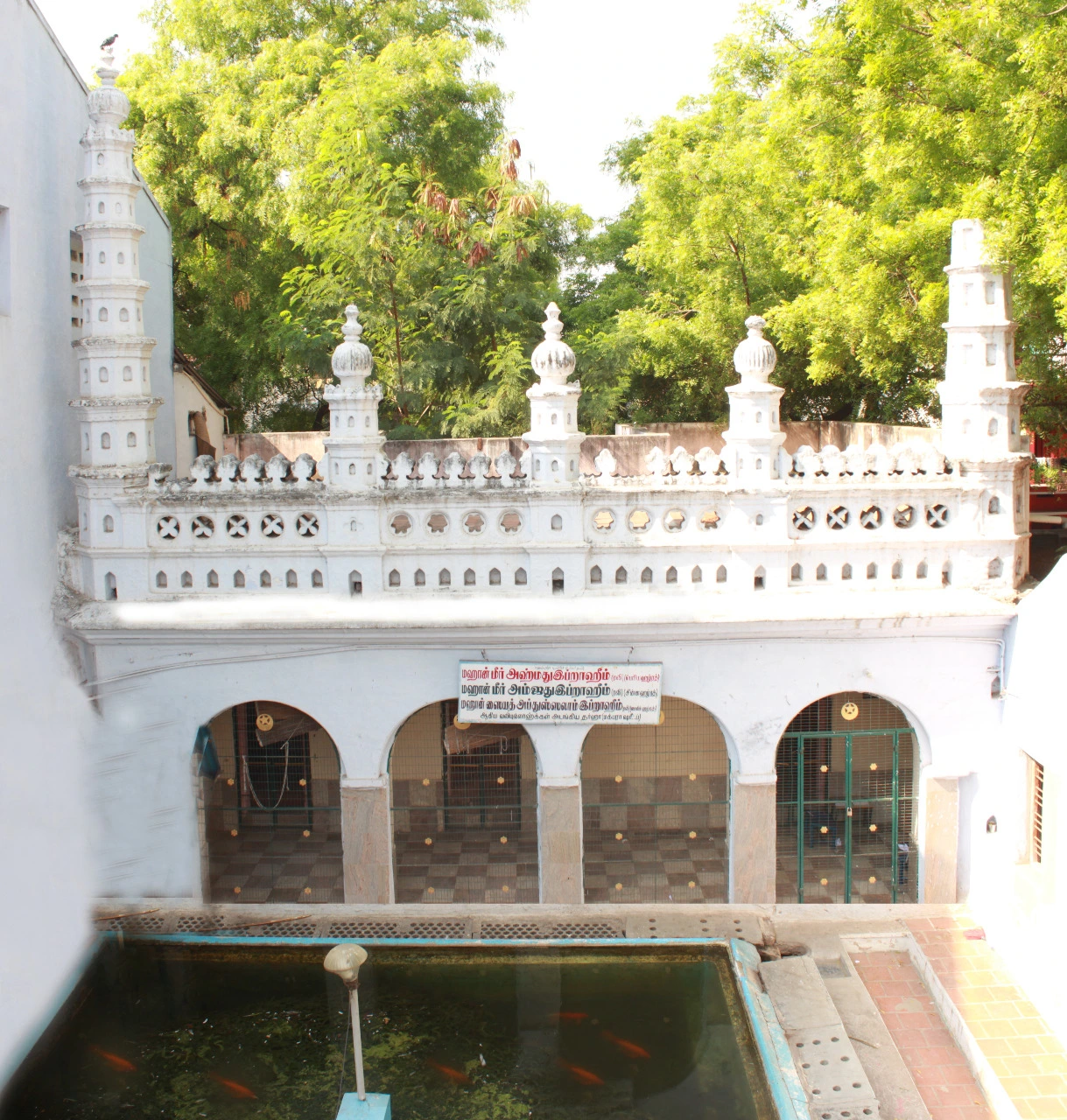 A whitewashed shrine building in South Indian style, with tiered pinnacle towers at its corners, a pierced parapet, and four arched openings closed with grilles beneath a signboard in Tamil script, with a green water tank in the foreground