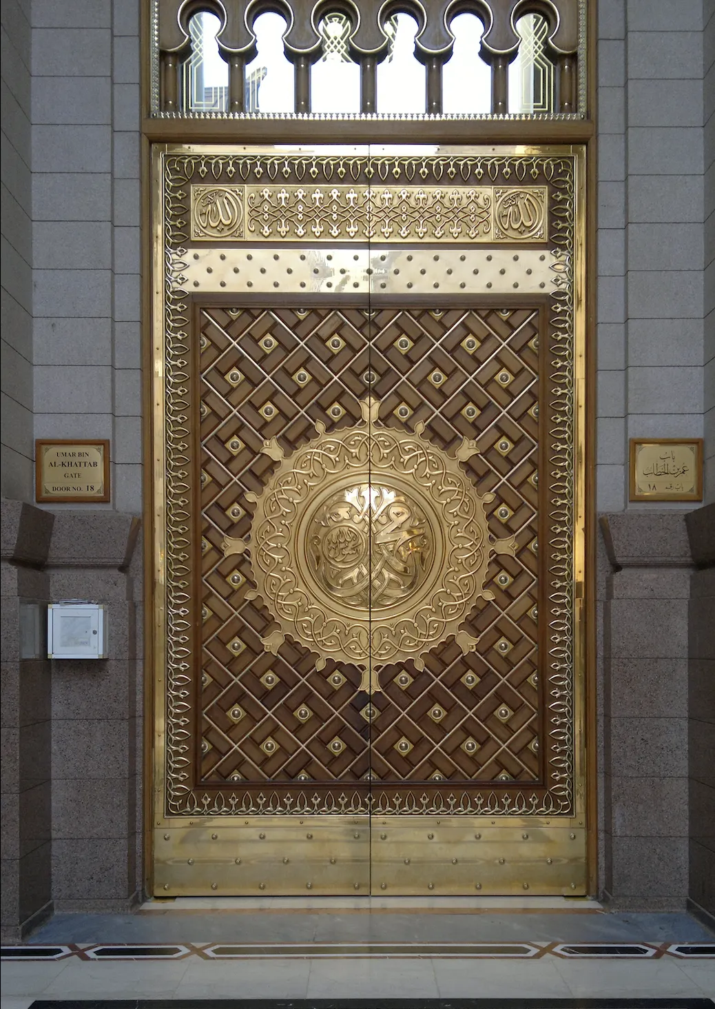 A tall double gate of brass and wood set in stone jambs, the leaves worked in a diagonal lattice around a large circular medallion of interlaced Arabic calligraphy giving the name of the Prophet Muhammad, with arabesque brass borders, the word Allah in the two upper corner panels, a pierced brass transom above and an inlaid marble threshold below; small plaques to either side name the gate in English and Arabic