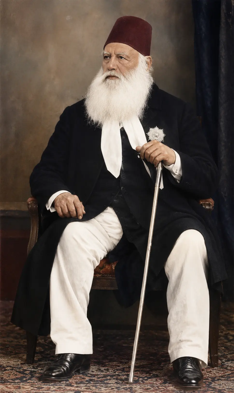 AI-generated impression: Studio portrait of an elderly man with a full white beard seated in a wooden armchair on a patterned carpet, wearing a dark red fez, a long dark coat over a dark waistcoat with a white neckcloth at the throat, white trousers and black shoes, a single silver breast star on his left lapel, his right hand resting on a slim silver-topped cane