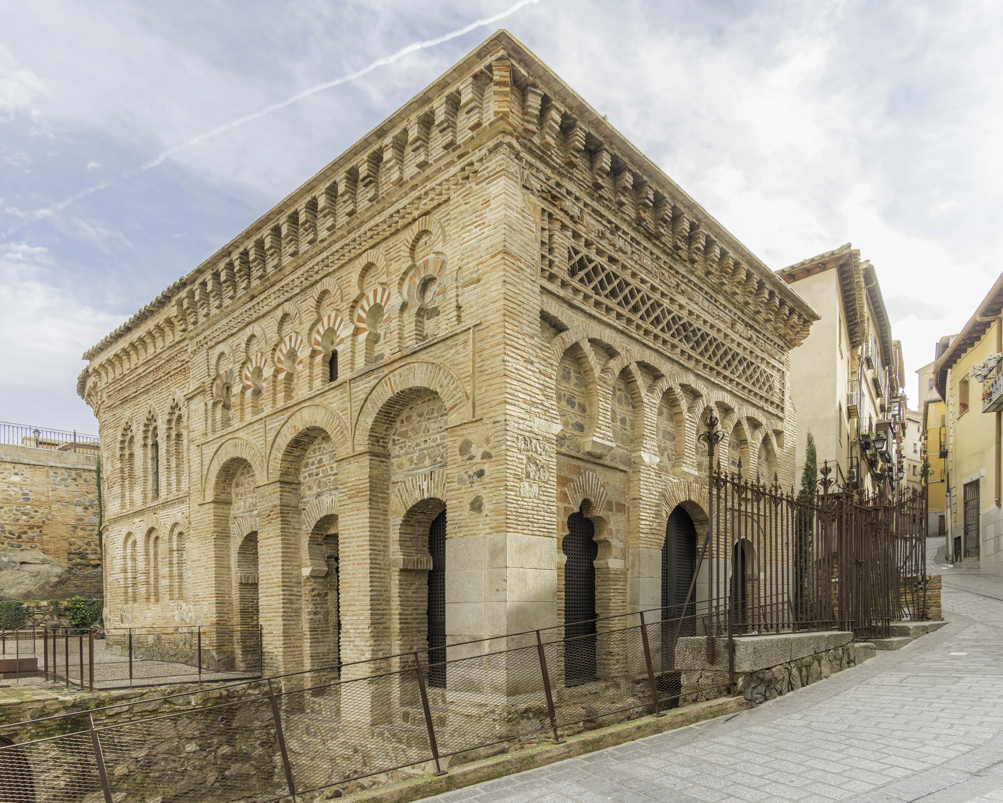 The corner of a small brick building with two tiers of horseshoe and multifoil blind arcading in an interlaced pattern, iron railings enclosing it beside a street